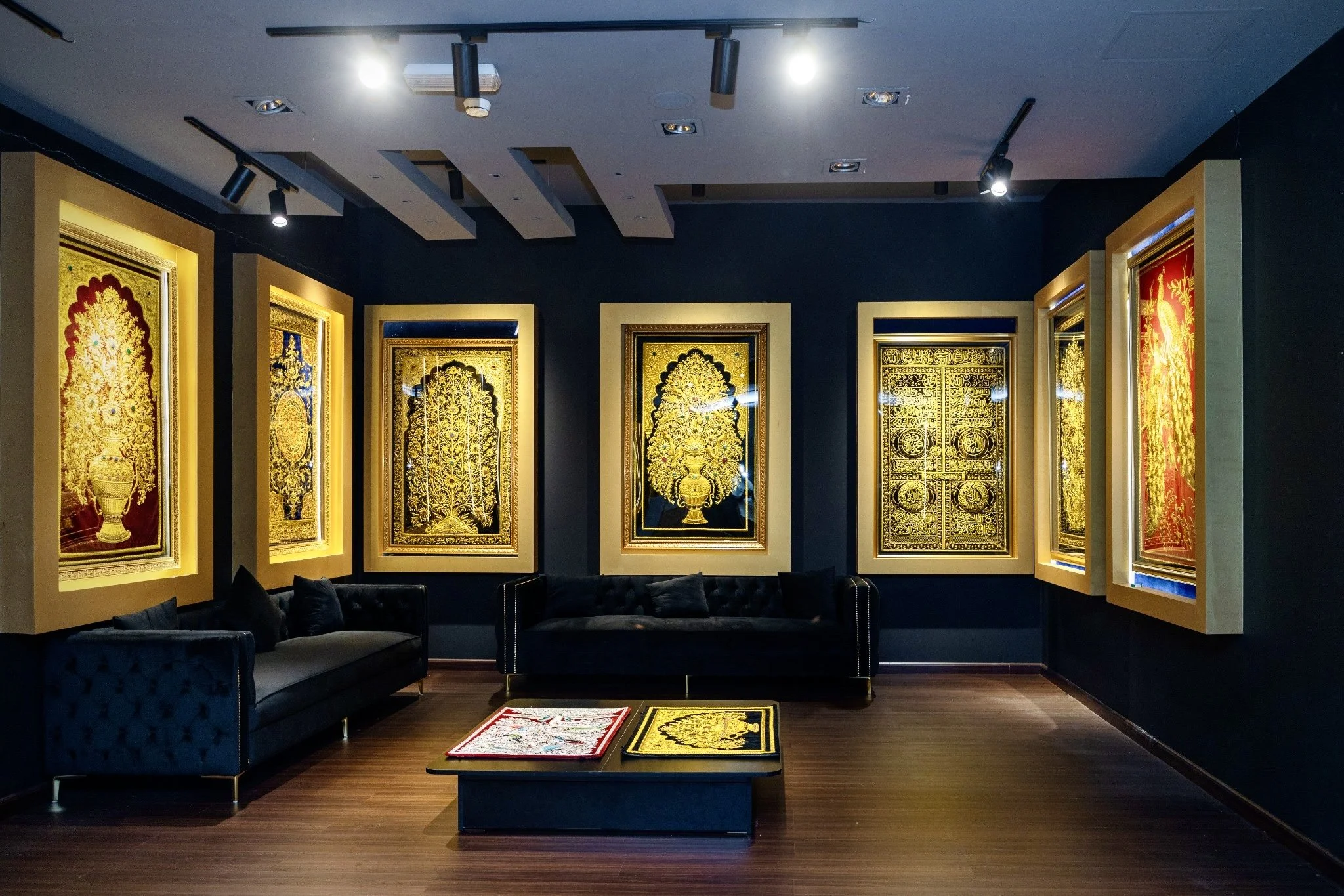 An art gallery or museum room featuring illuminated framed gold and red artwork with intricate designs hanging on dark walls, a black sofa with throw pillows, and a small table with colorful and black gold framed items on a wooden floor.
