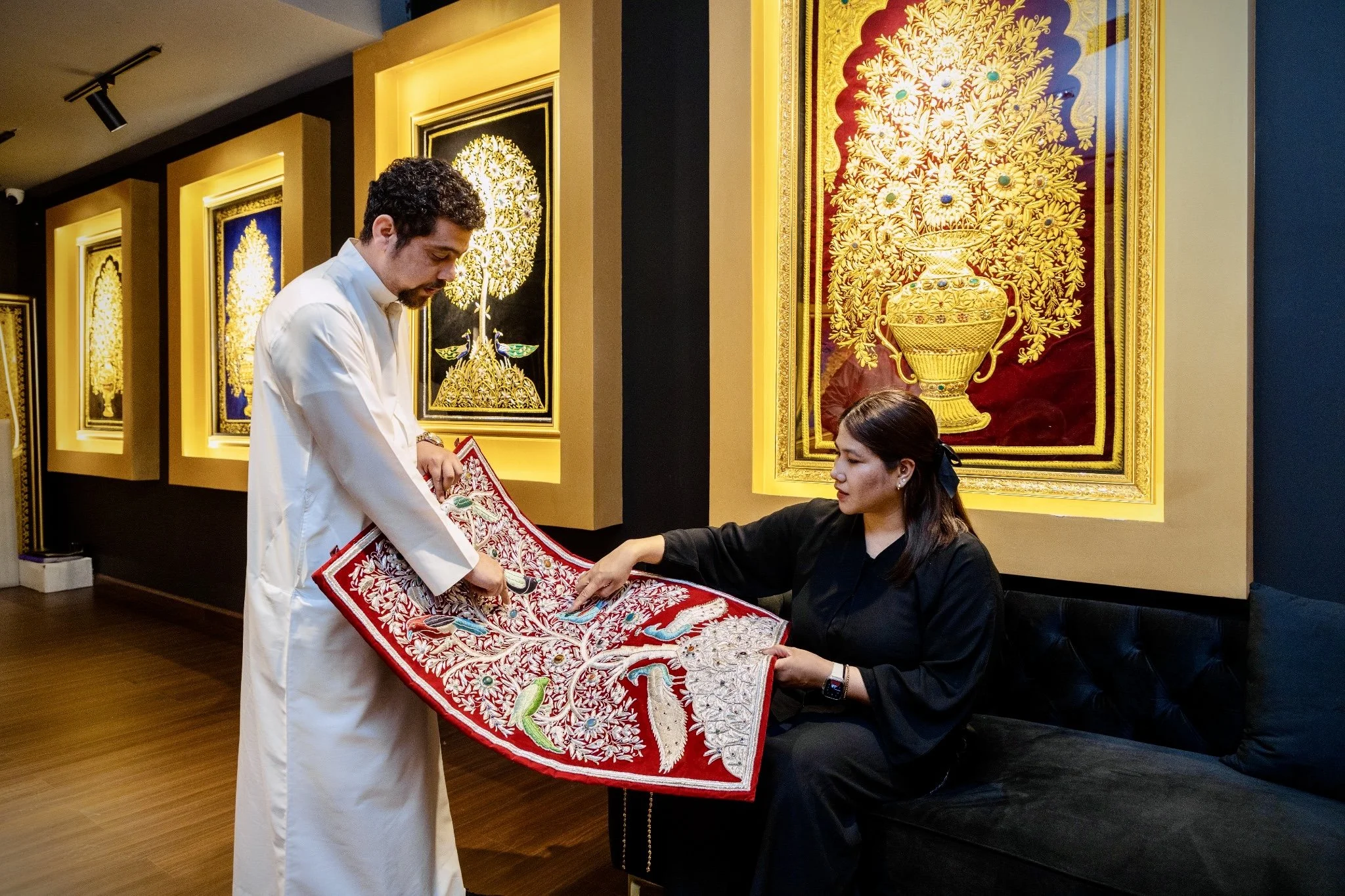 A man dressed in white traditional attire presenting an embroidered red and white cloth to a seated woman in a black outfit, inside an art gallery with vibrant framed artwork on the wall.