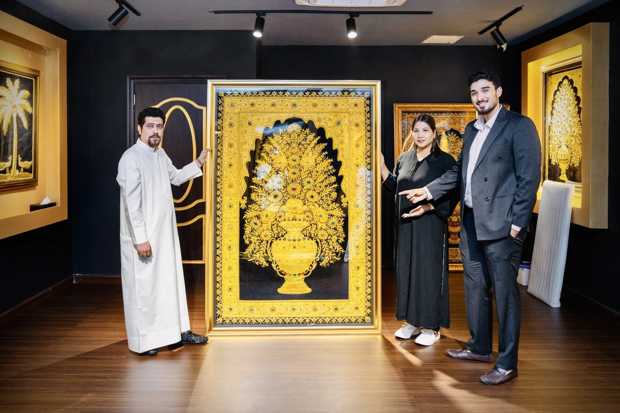 Four people standing in front of an ornate gold and black artwork featuring a floral arrangement and a vase, inside an art gallery. The group includes three men and one woman, all dressed formally or semi-formally.