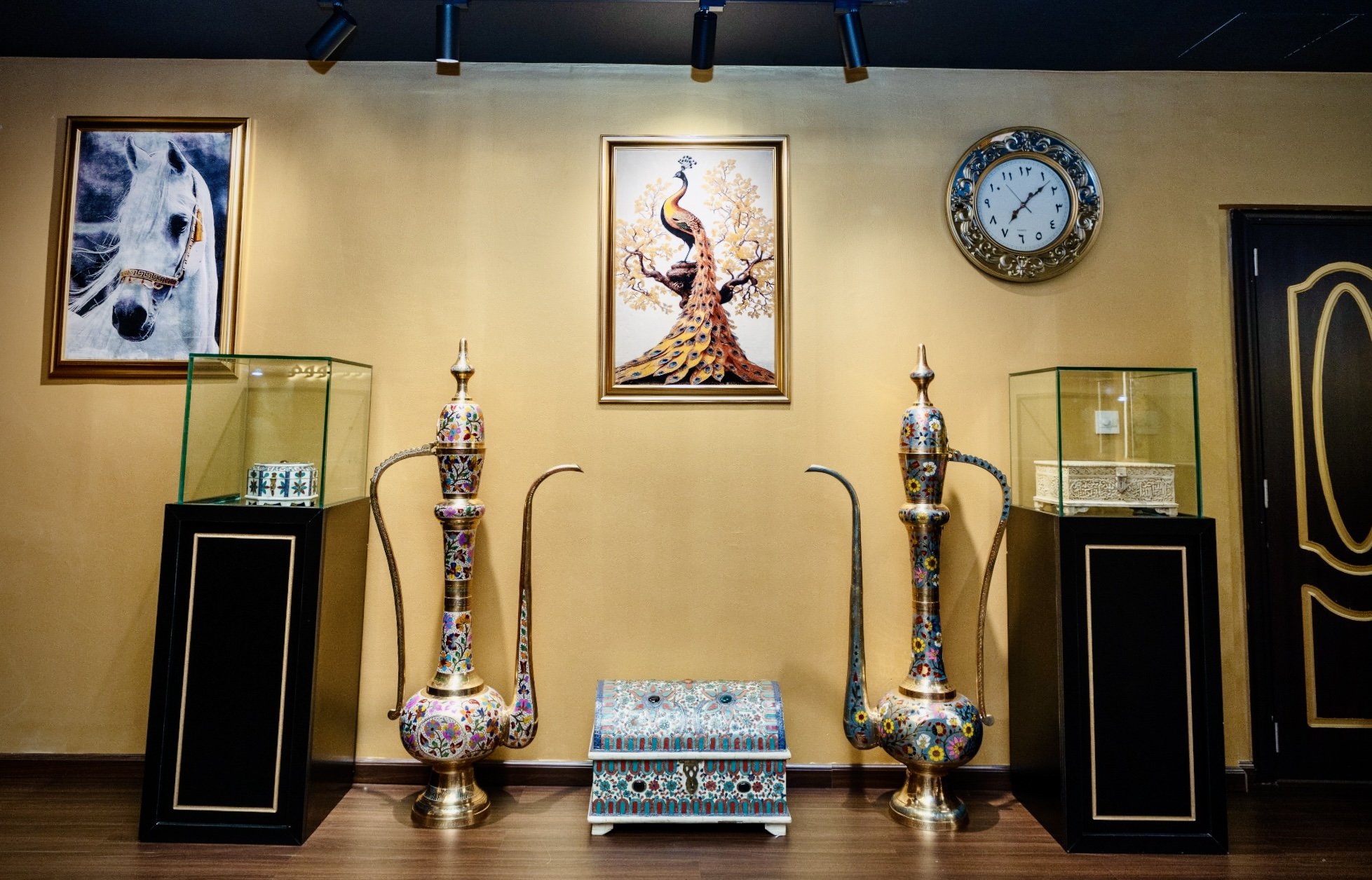 Display of decorative objects including three tall, colorful vases with intricate patterns, a small box, and a table with floral motifs. Two framed pictures of a white horse and a peacock are hanging on the yellow wall. A round ornate clock is mounte