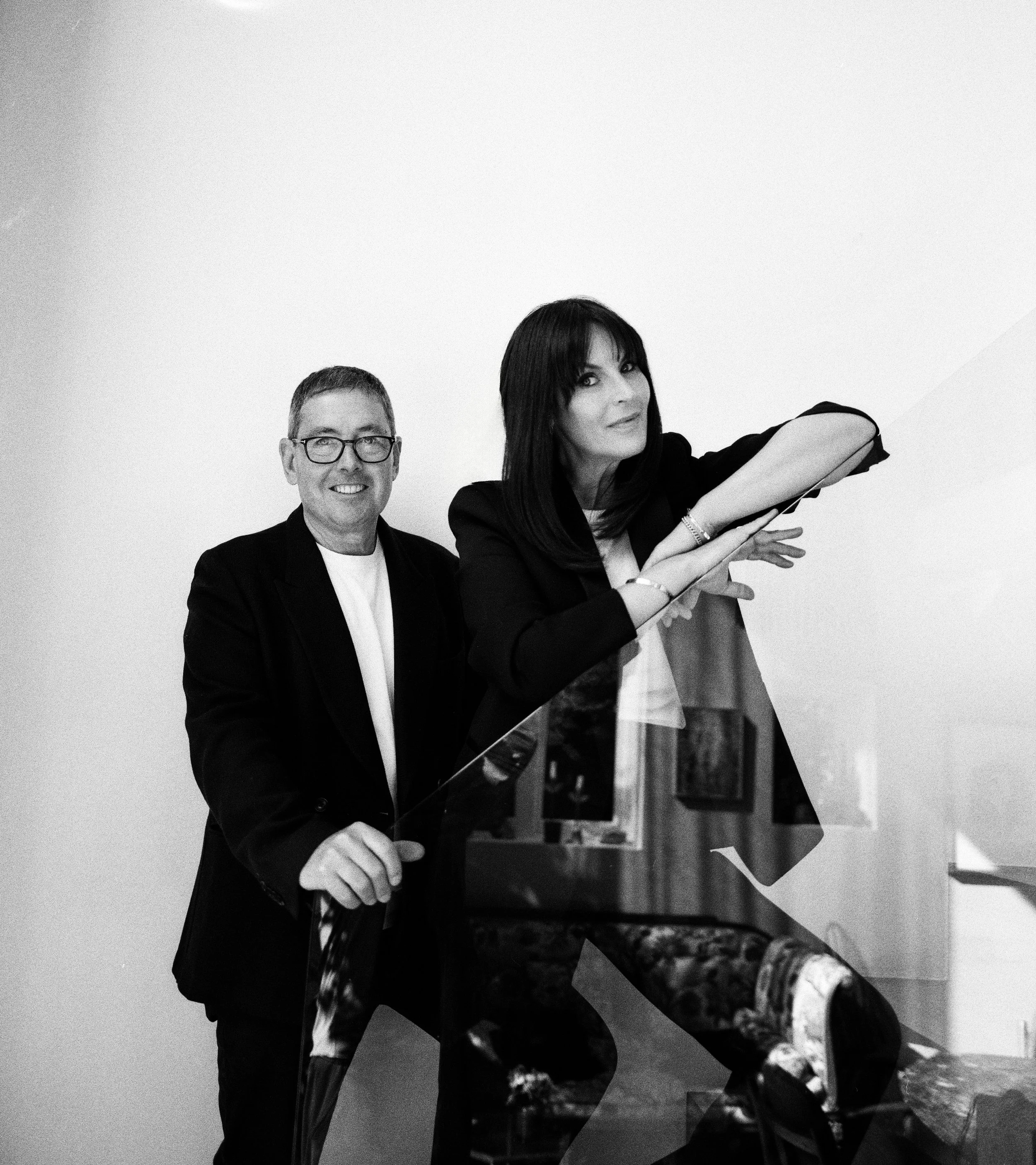 Black and white photo of a man and woman standing behind a glass panel. The woman is leaning on it with her arm, and the man is holding the edge of the panel. The woman has dark hair and is wearing a blazer, while the man is wearing glasses and a suit. The background is plain, and the setting appears to be indoors.