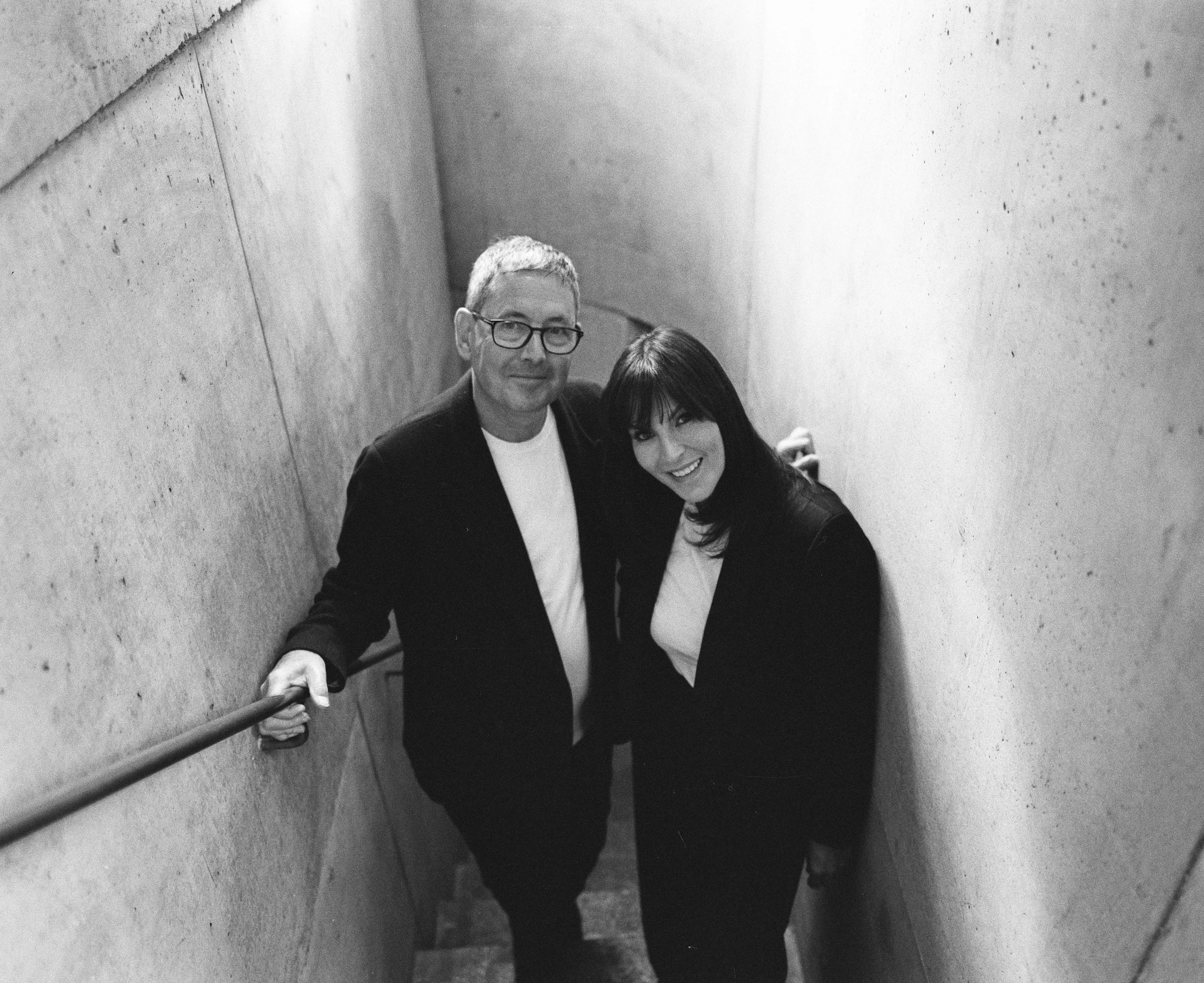 A black and white photo of a man and woman smiling, standing on a narrow stairwell with concrete walls. The man has short hair, glasses, and is wearing a suit, while the woman has long dark hair and is dressed in a dark jacket and white top.