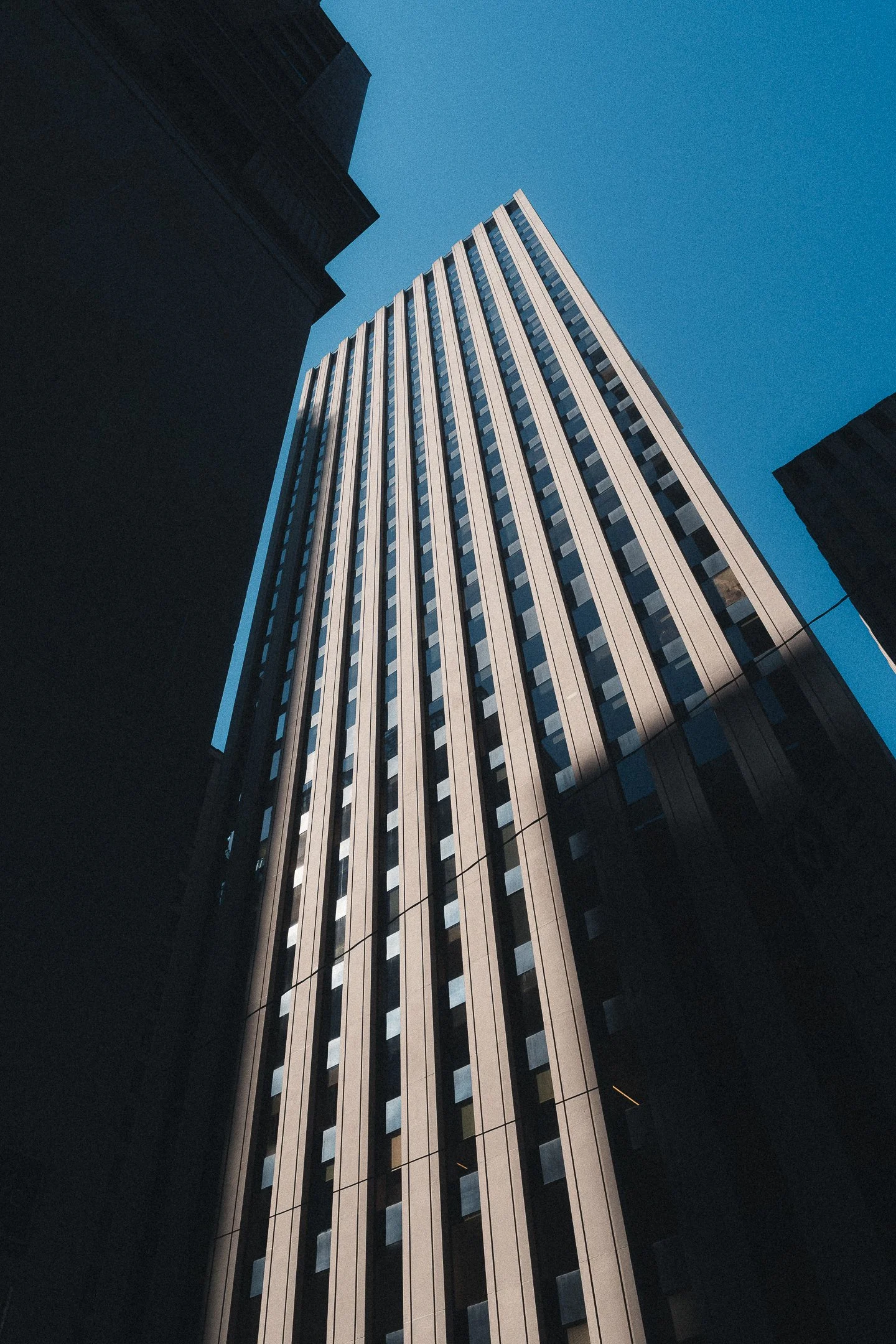 Looking up at a tall skyscraper with vertical lines and windows, surrounded by other tall buildings under a clear blue sky.