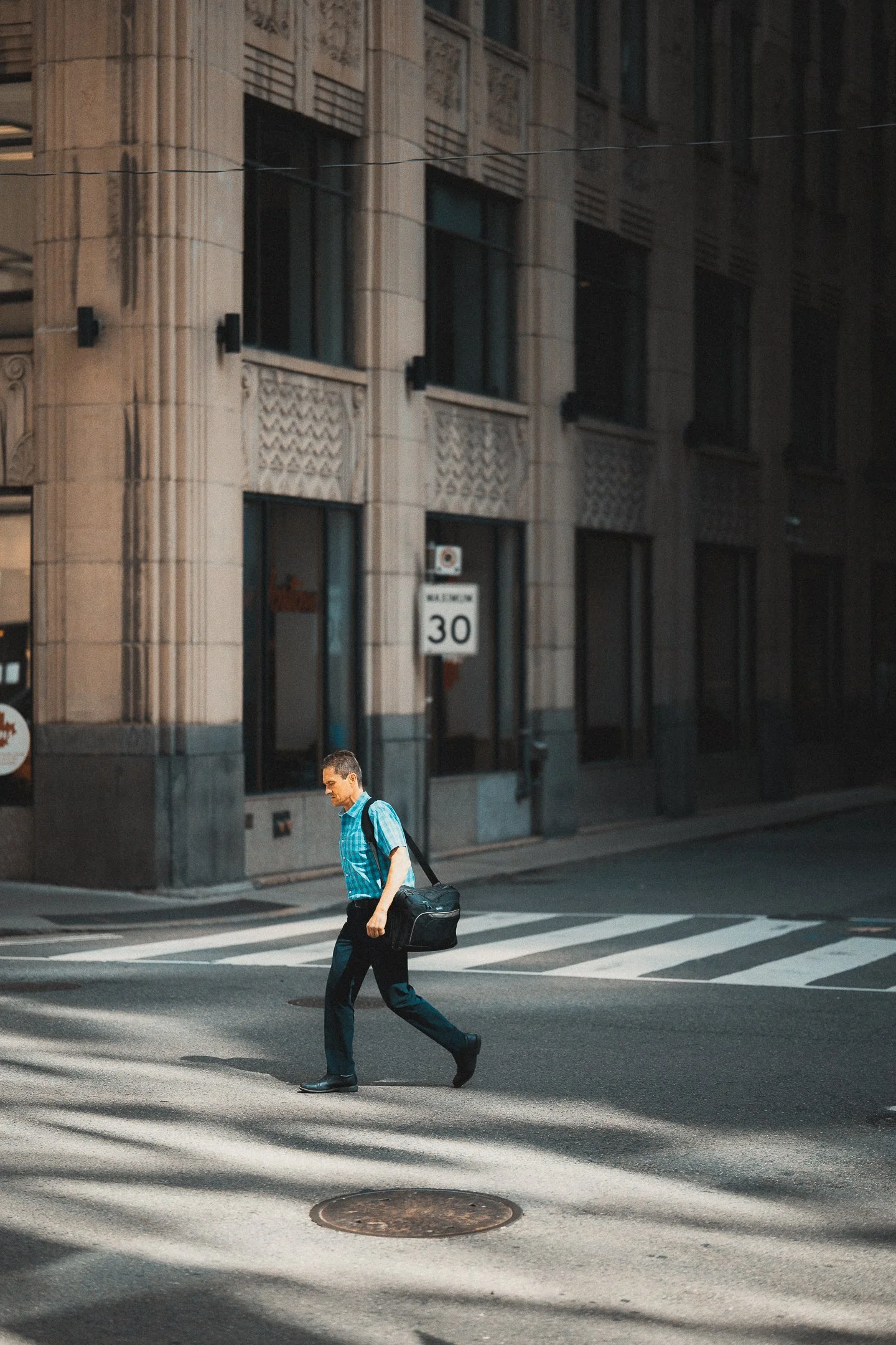 A man in a blue checkered shirt walking across a city crosswalk with a black shoulder bag. The background features a tall, ornate building with tall windows and a traffic sign displaying a speed limit of 30.