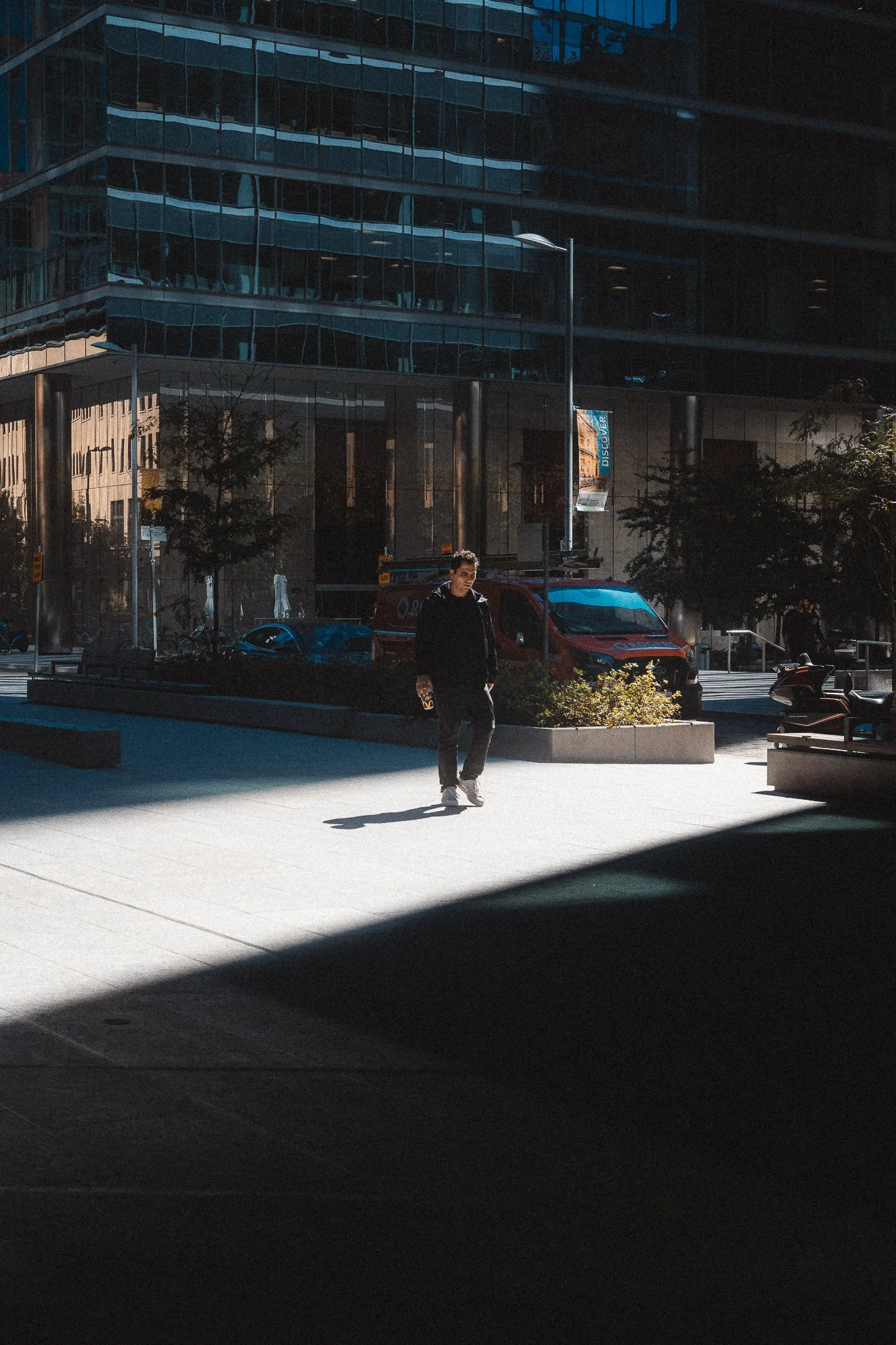 A man walking on a city sidewalk in front of a modern glass building, with a red service van parked nearby, and shadows cast across the scene.