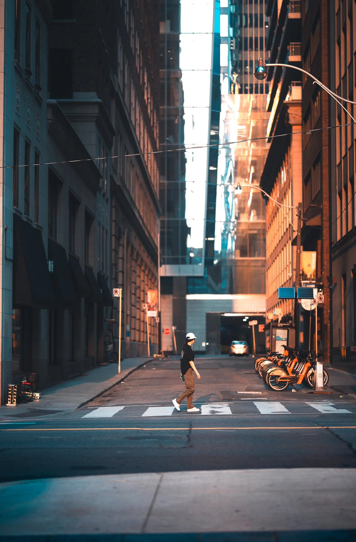 A person walking on a crosswalk in a city street at sunset, with tall buildings on either side and a row of orange bikes parked on the right.