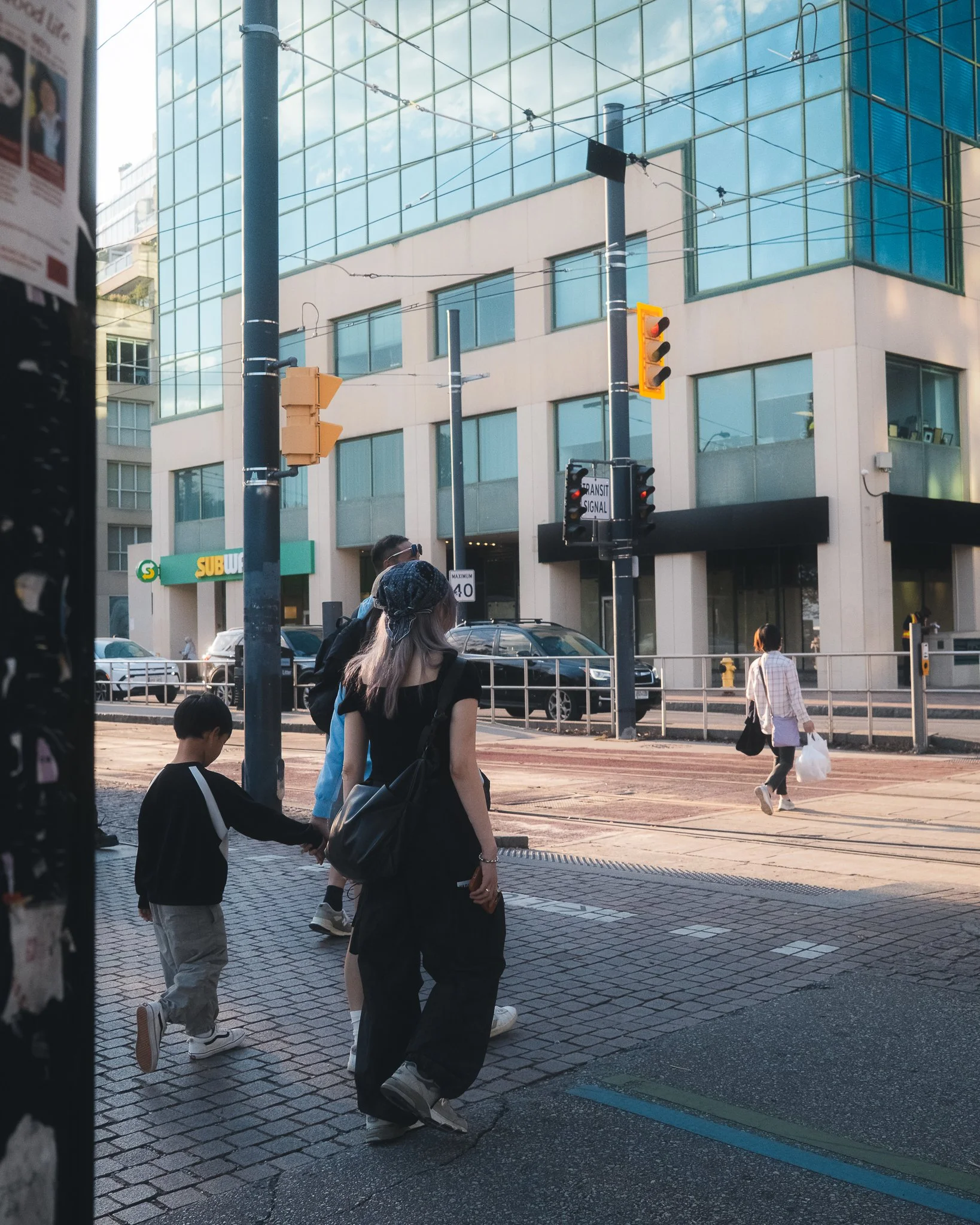 People crossing the street in an urban area with modern glass buildings, traffic signals, and a Subway restaurant sign.