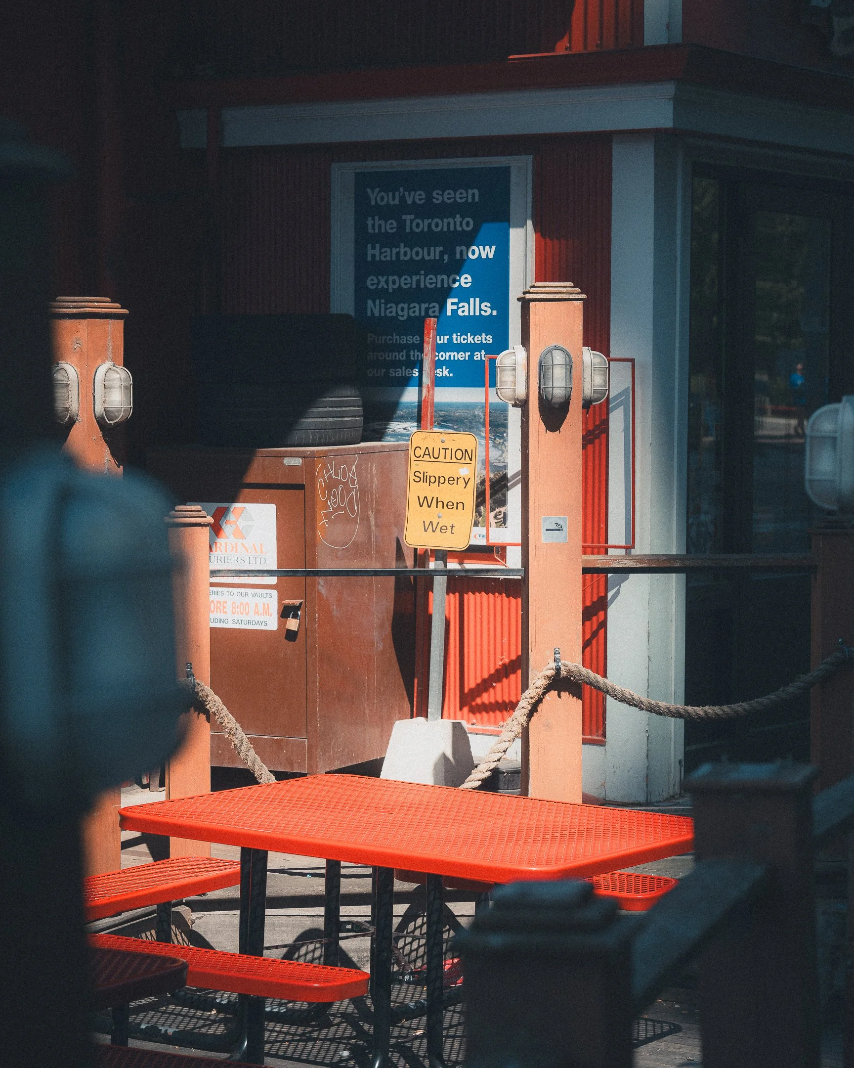 Outdoor seating area with a bright orange table and benches, a caution sign warning slippery when wet, and a poster promoting experiences at Niagara Falls behind a railing with ropes, in front of a building with red siding and a large window.