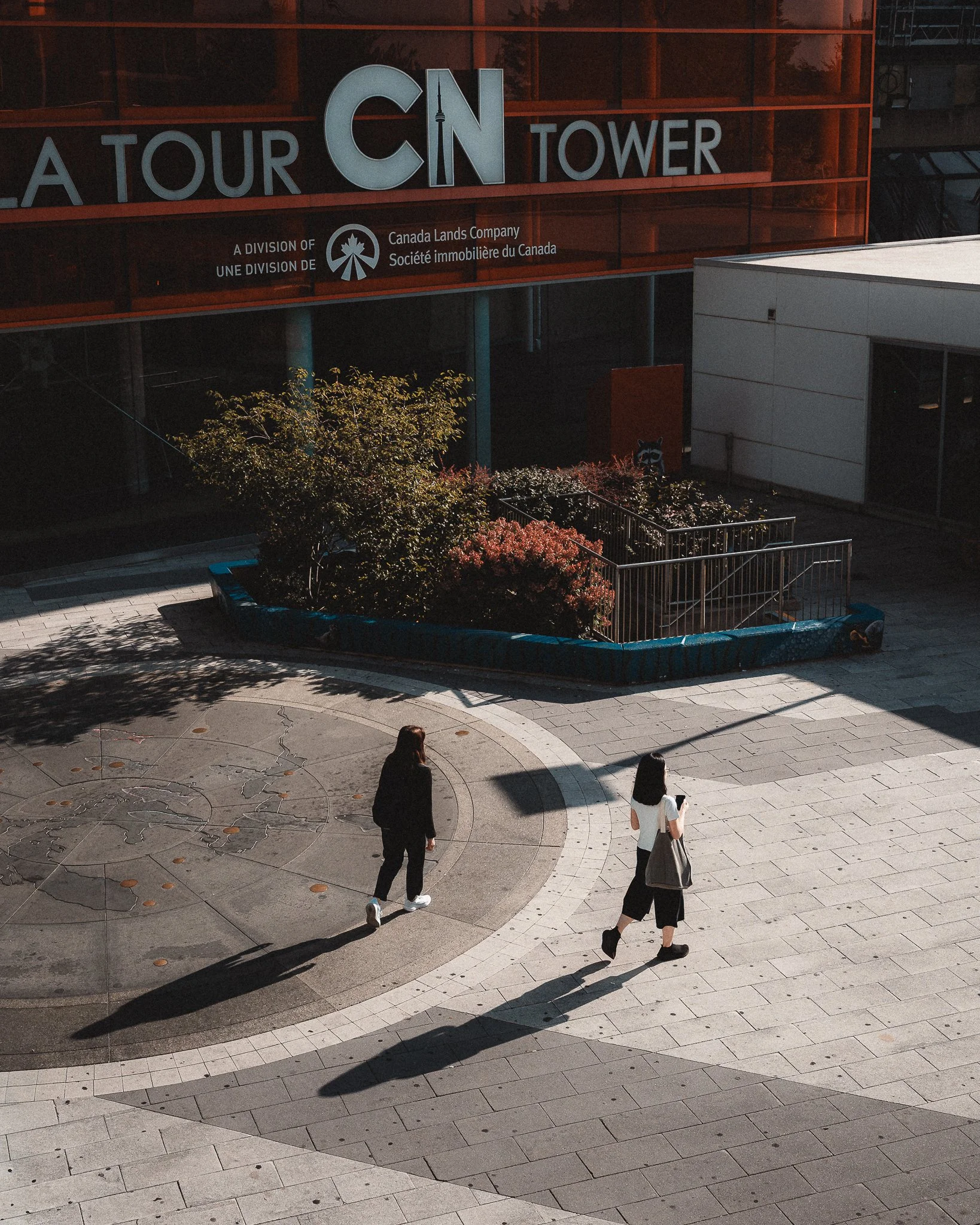 Two women walking outside a tower with a large sign that reads 'CN Tower' and a small garden with trees and bushes.