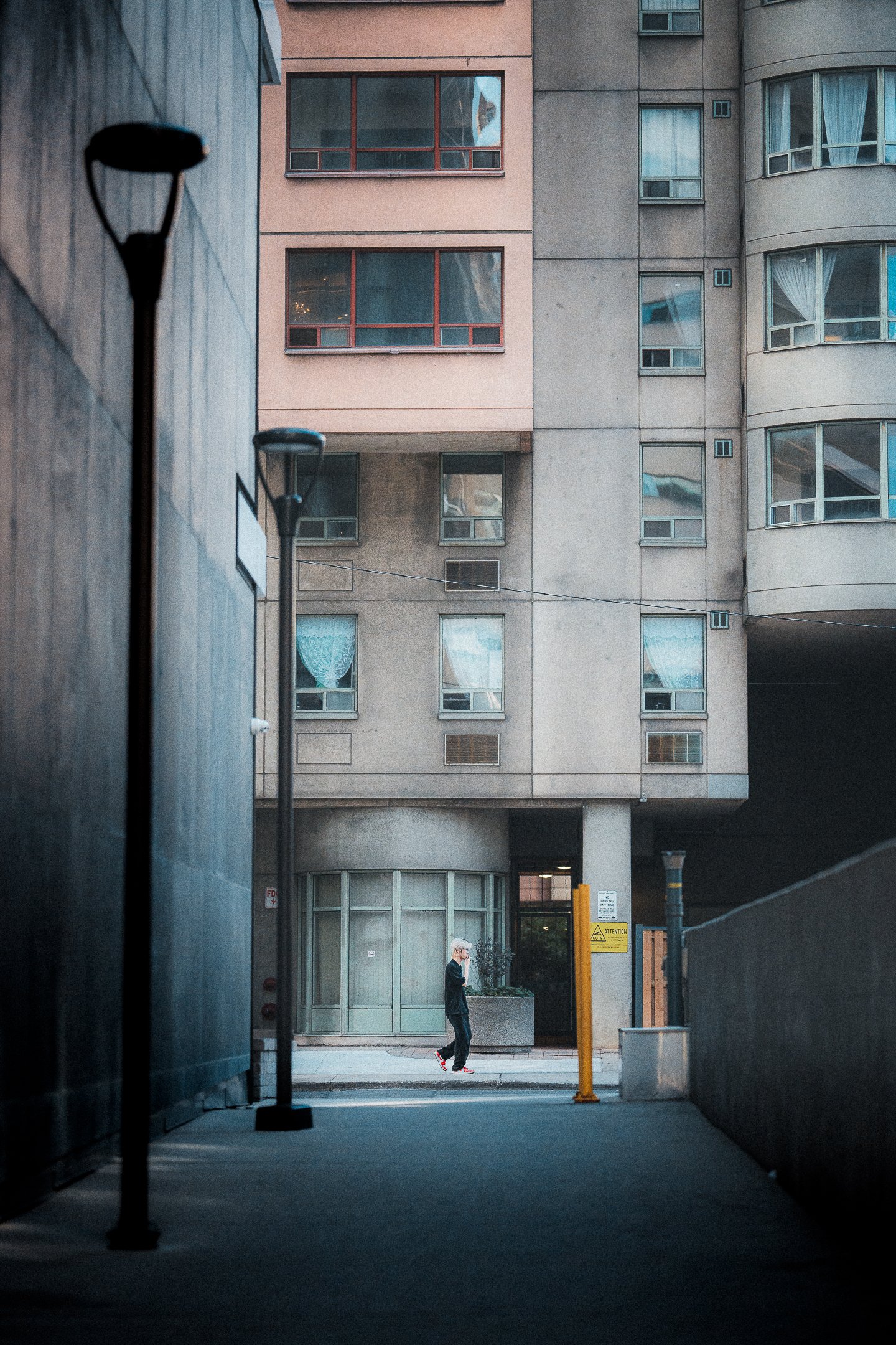 A person walking and talking on a cell phone along a city sidewalk in front of a tall, modern apartment building, with lamp posts and parking barriers in the foreground.