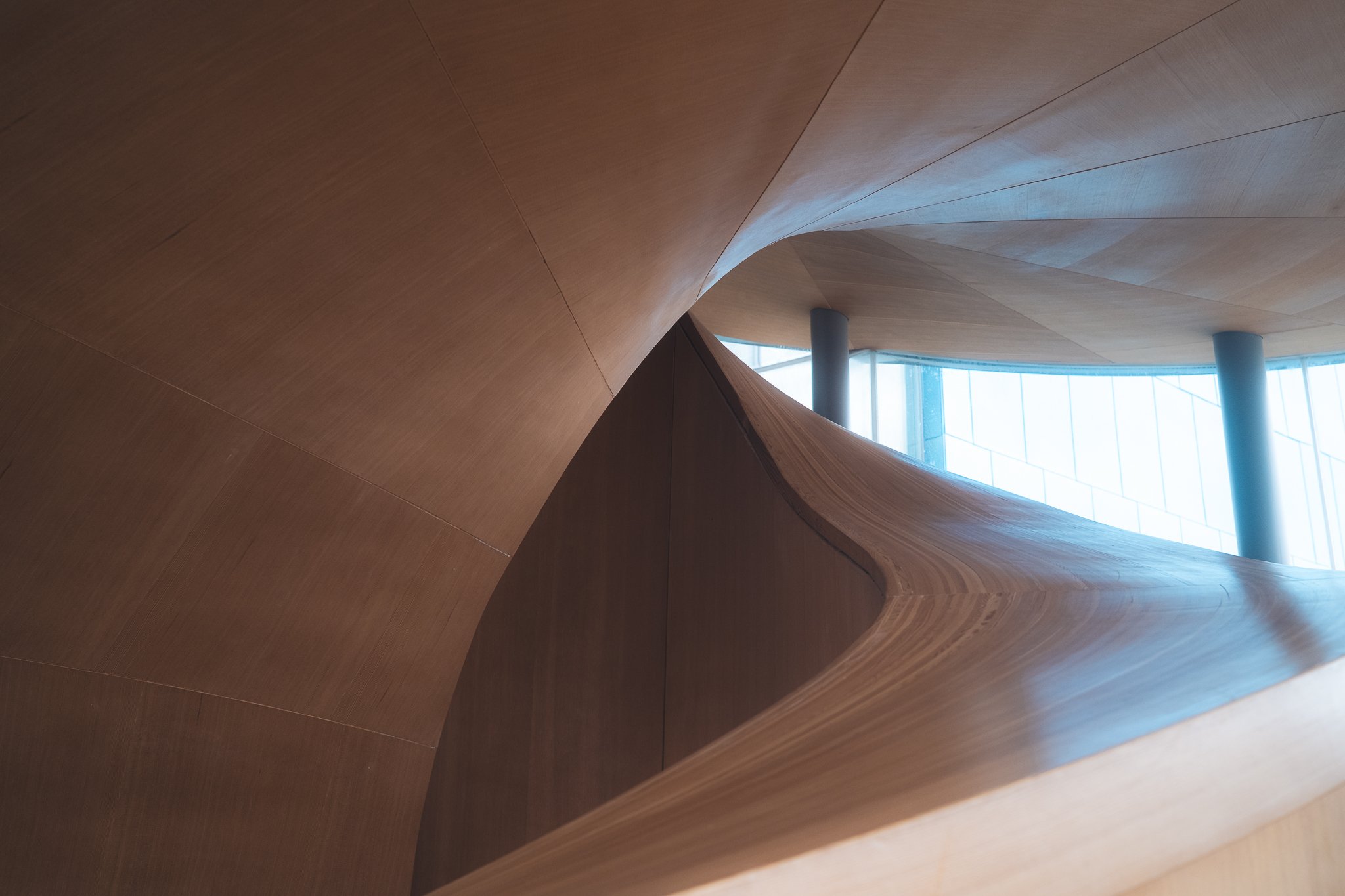 Interior view of a modern wooden staircase with smooth curves and a glass wall in the background.