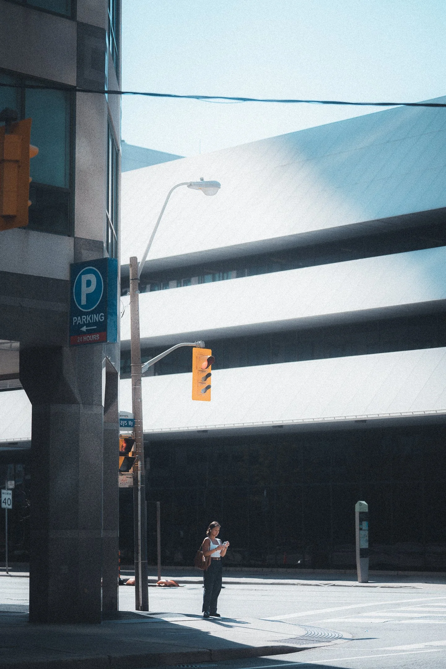 A woman standing on a city street corner looking at her phone in front of a parking garage, with modern buildings in the background and a red traffic light overhead.