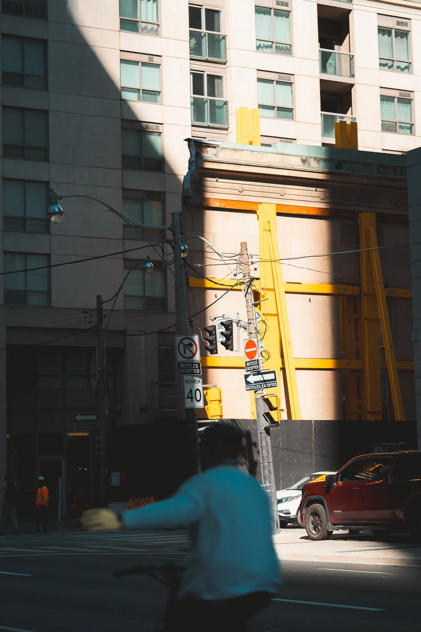 A street scene in front of a building with construction scaffolding. A cyclist is in the foreground, partially blurred, with traffic signals and street signs visible, including no left turn and maximum speed of 40 mph signs. Two people in orange vest