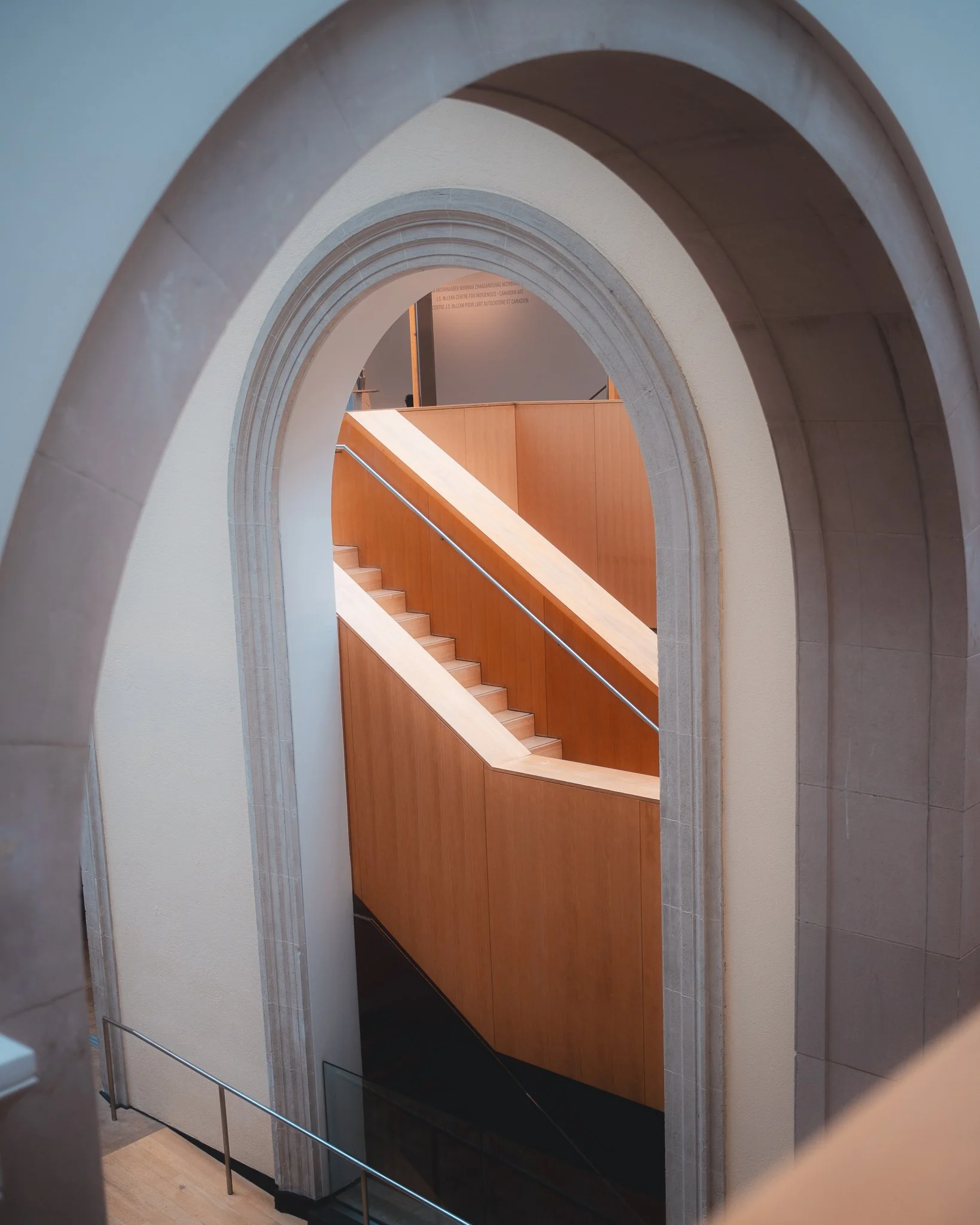 Interior view of a staircase seen through decorative archways with beige walls, wooden paneling, and silver handrails.