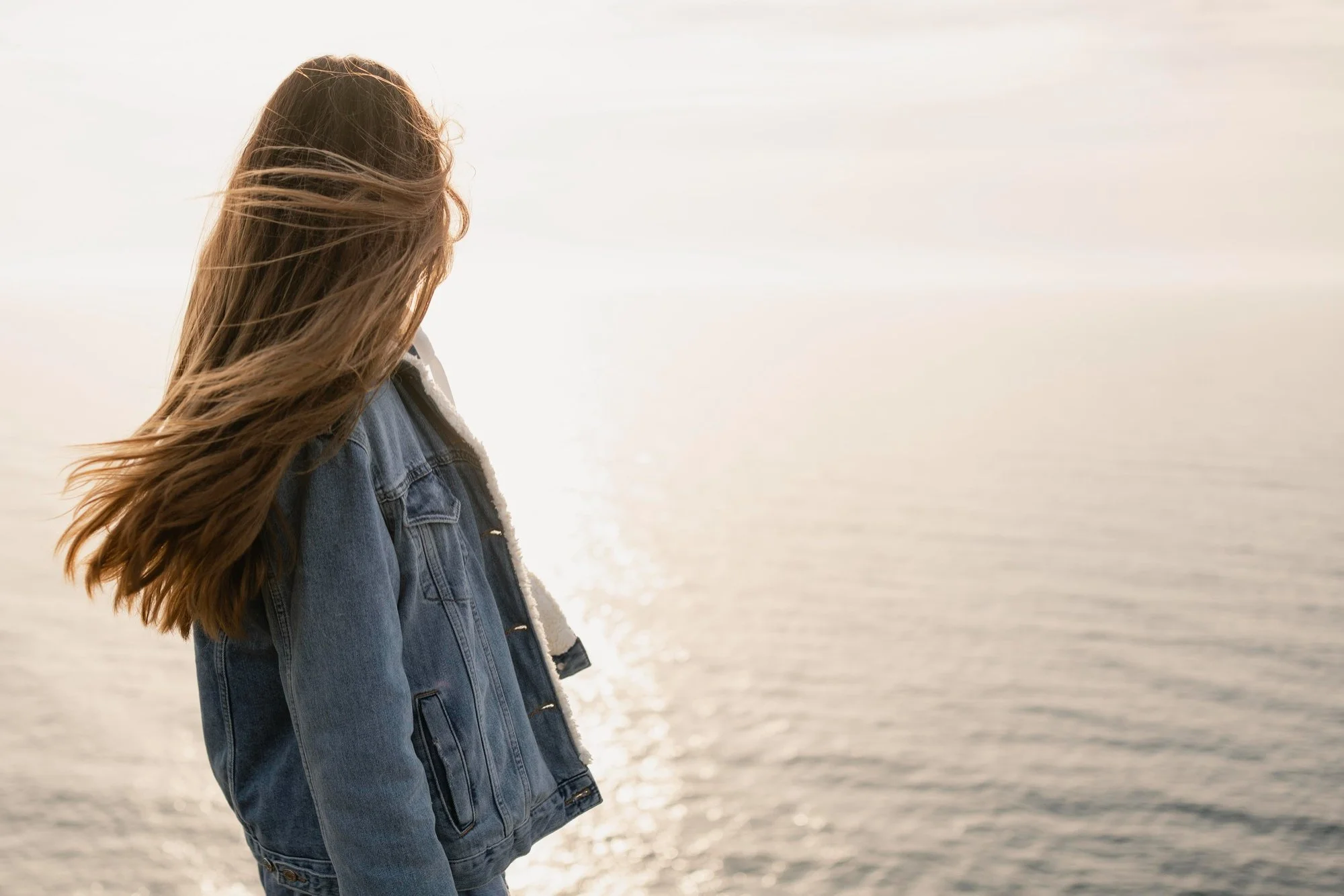Junge Frau mit langen hair, die an einem Strand bei Sonnenuntergang steht, trägt eine Jeansjacke.