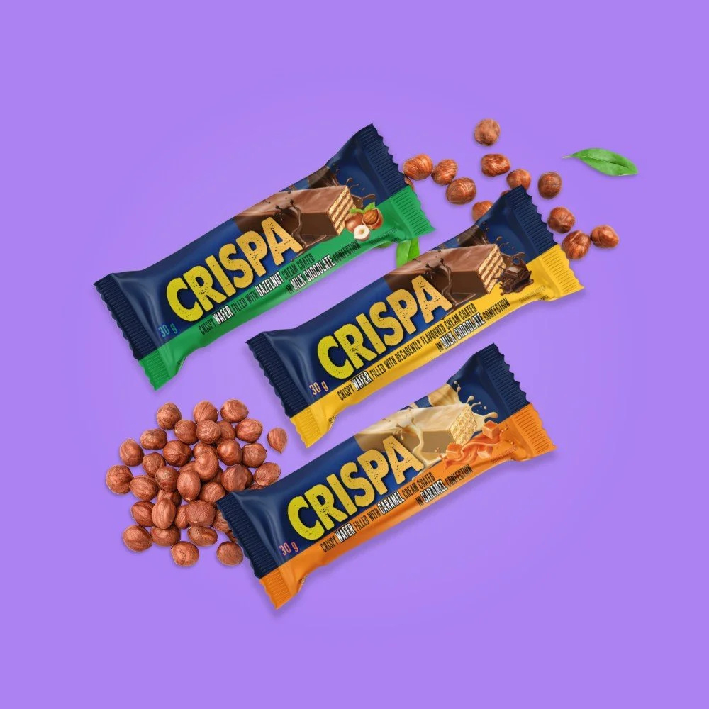 Three Crispa snack bars, one with a green label, one with a yellow label, and one with an orange label, are displayed on a purple background with scattered hazelnuts and a green leaf.