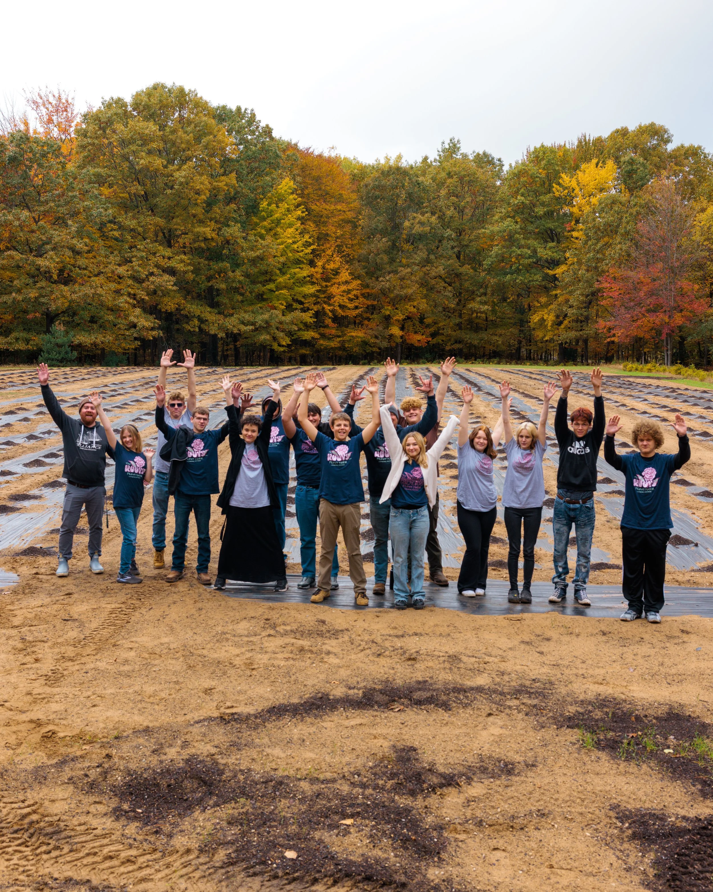 Group of people standing with hands raised in an outdoor farm or garden field surrounded by autumn trees