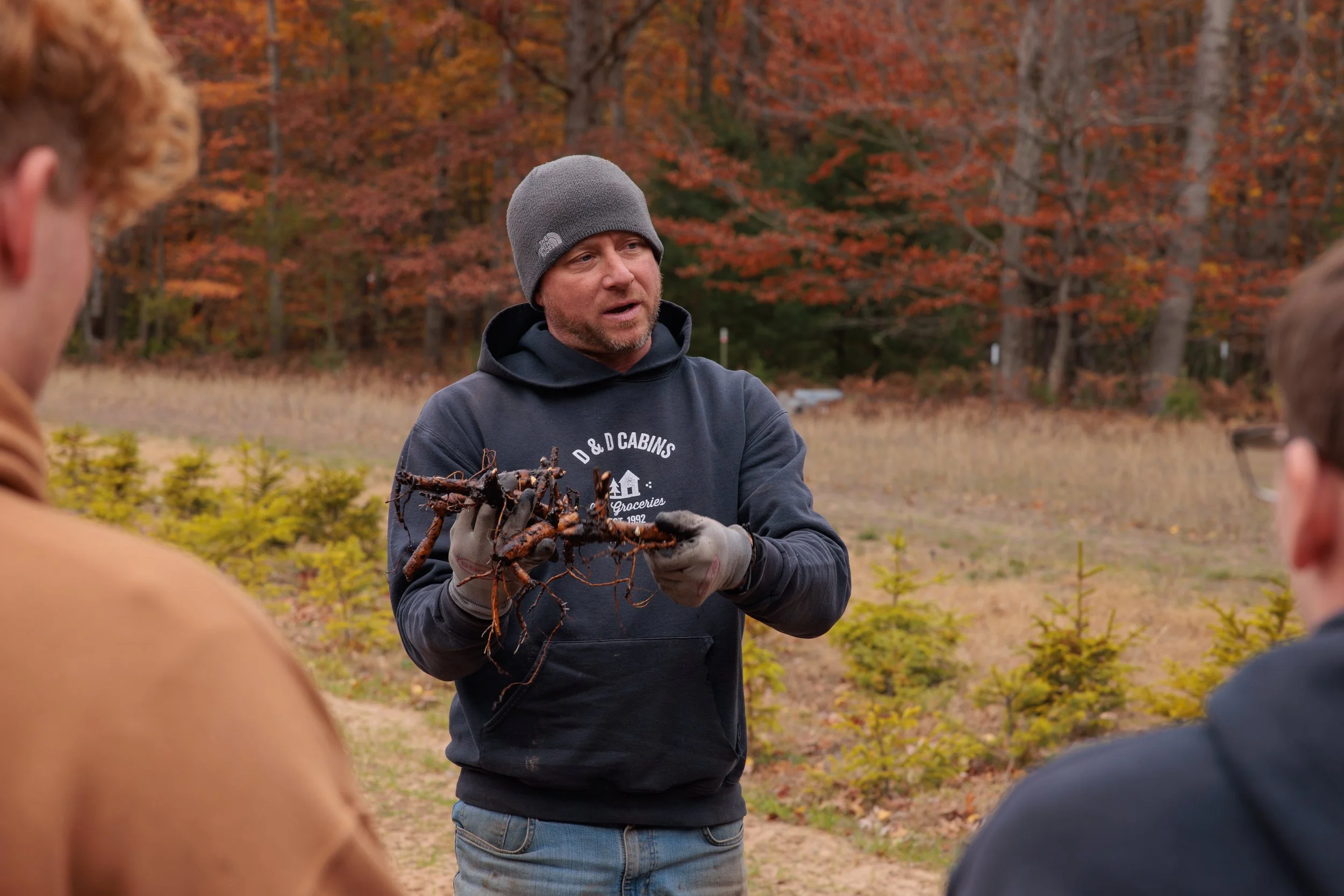 Man in black hoodie and gray beanie holding roots or branches, explaining to a group outdoors with autumn-colored trees in the background.
