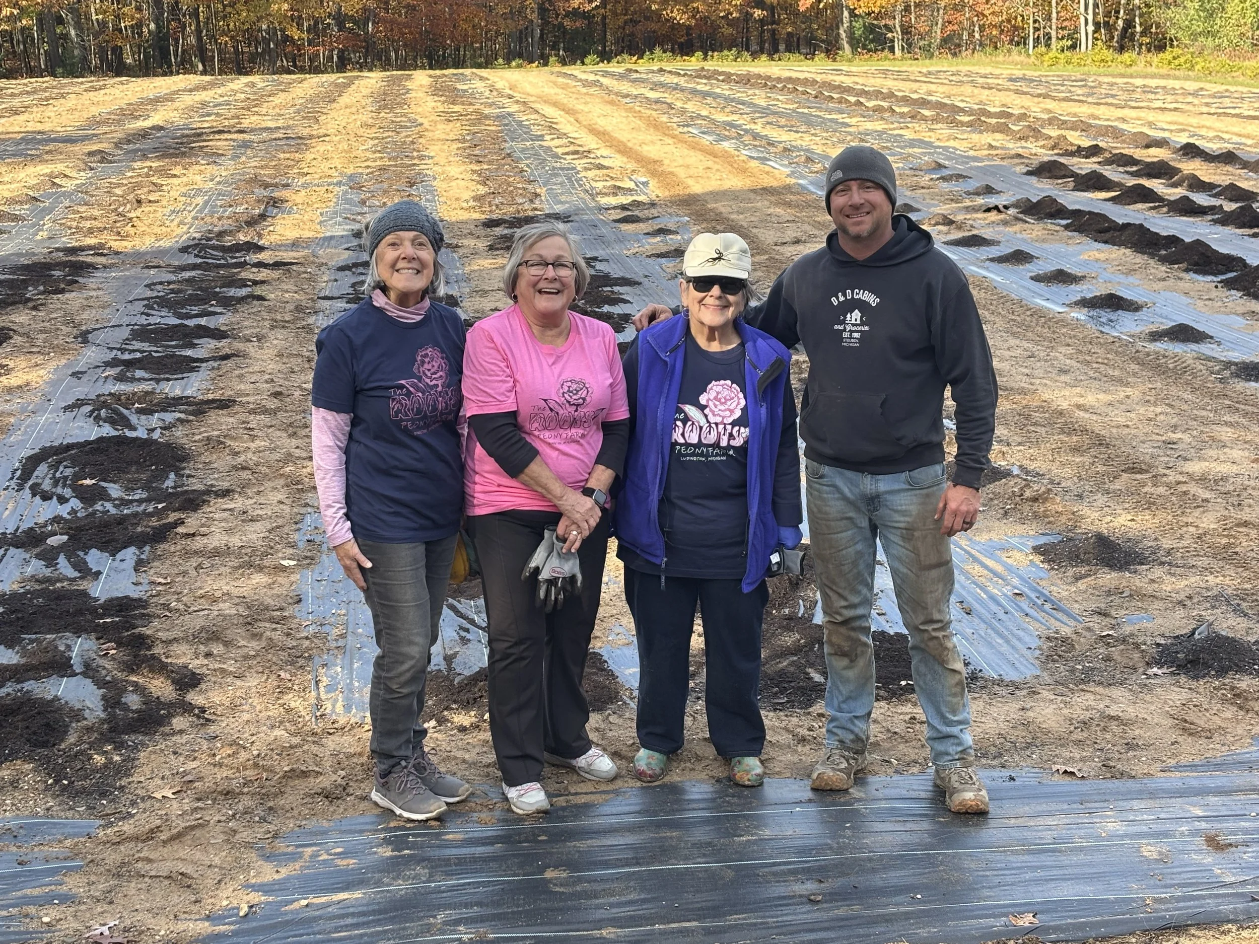 Four people standing in a farm field with plastic mulch and rows of soil, trees in the background, wearing casual outdoor clothing and smiling at the camera.
