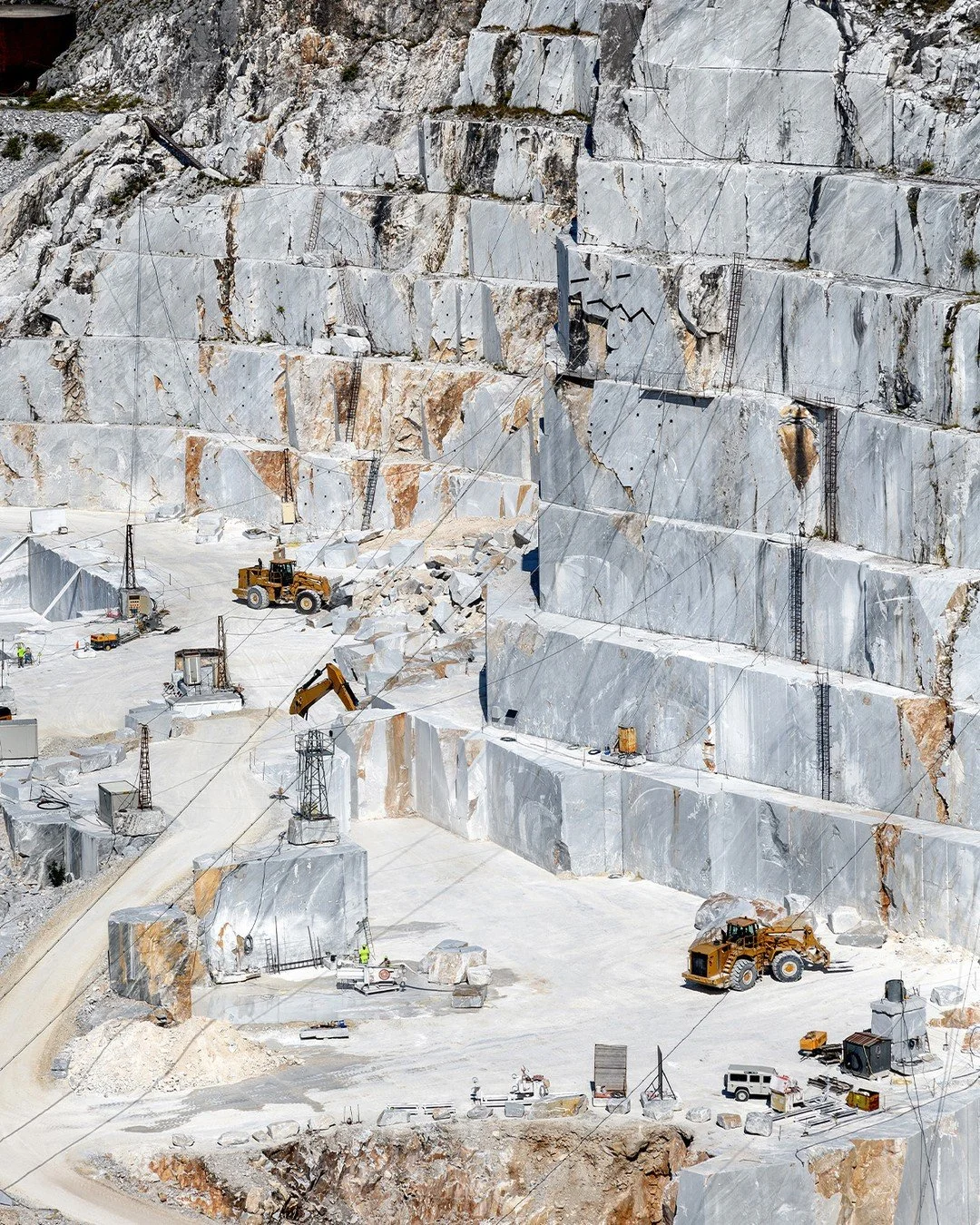 An elevated view of the terraced Carrara marble quarries, where centuries of craftsmanship and geology converge. Each step of this open-cast landscape reveals the monumental scale of extraction&mdash;towering walls of stone, precise cuts, and the con