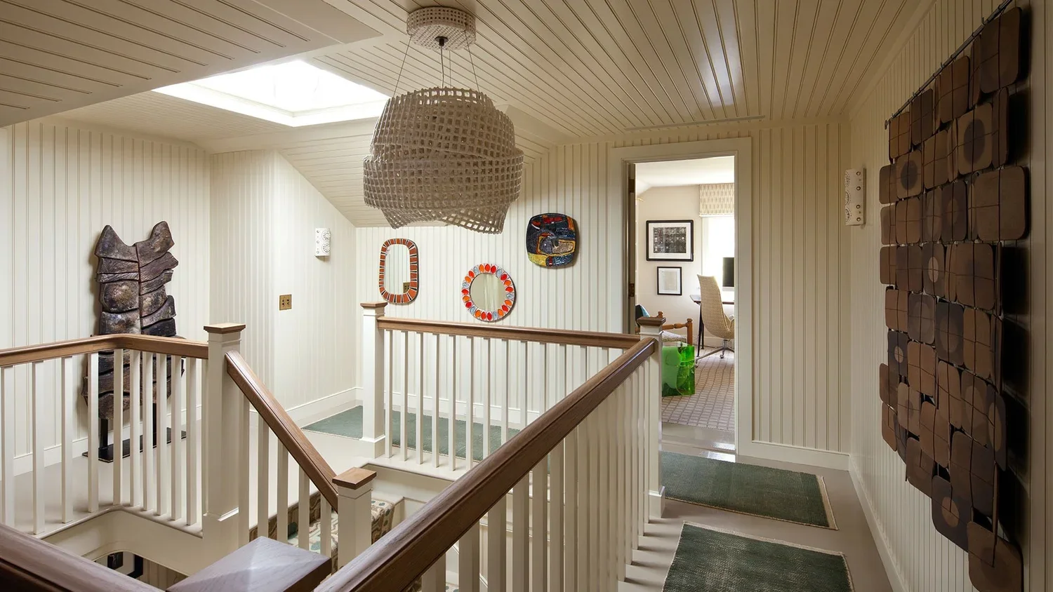 Interior view of a upper floor hallway with wooden railings, a ceiling skylight, and wall decorations. An open door leads to a workspace with a desk, chair, and artwork.