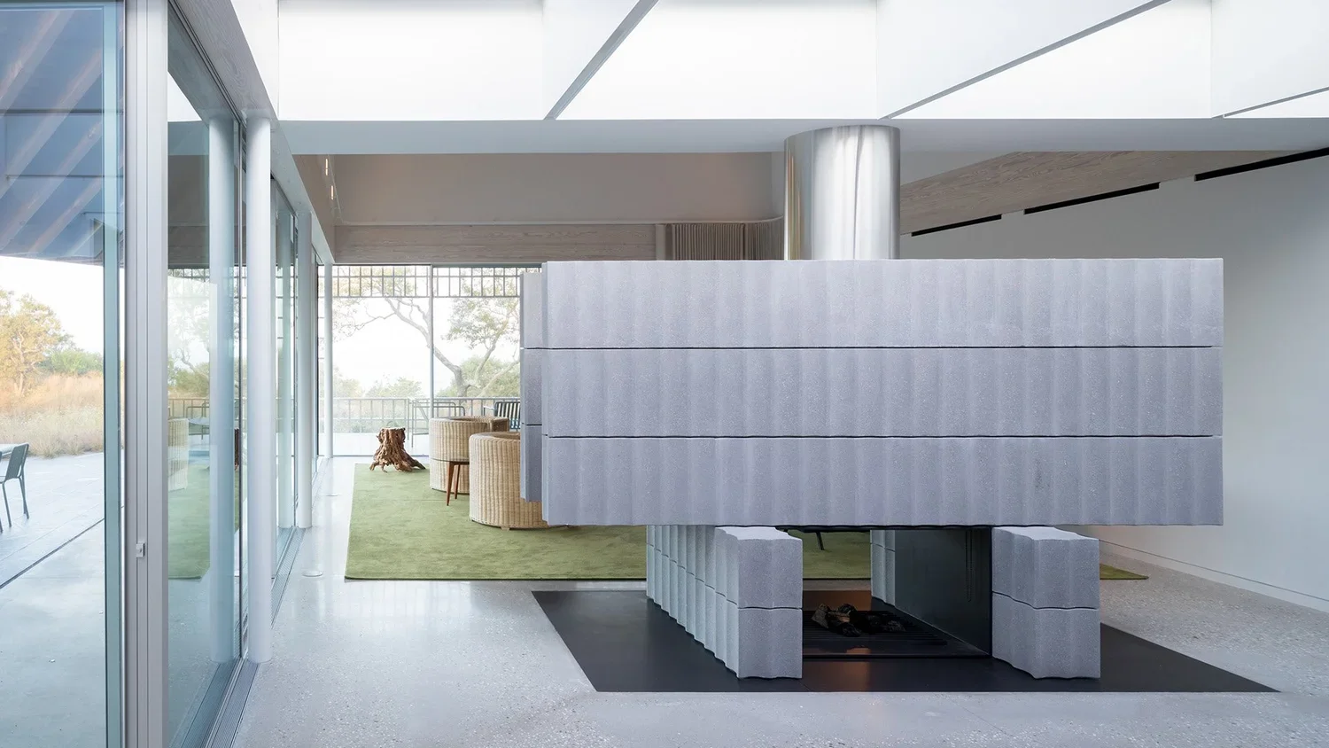 Modern interior with a large concrete fireplace, sitting area with beige chairs, green rug, and glass sliding doors leading to an outdoor area with trees.