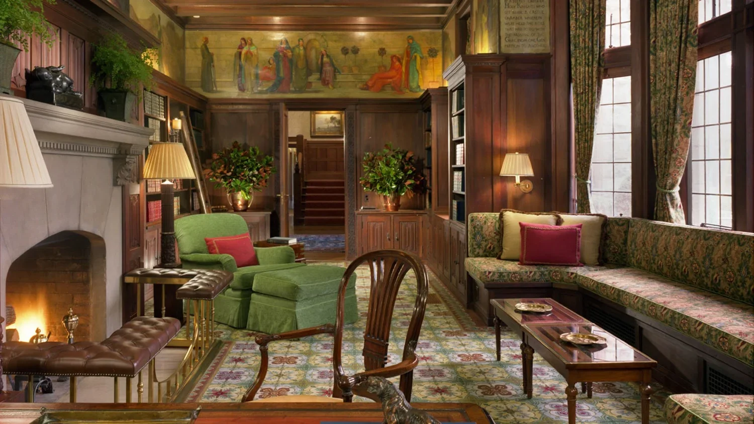 A cozy living room with wood-paneled walls and large windows with floral curtains, includes a green armchair with a red pillow, a floral-patterned built-in sofa with pillows, a fireplace, a wooden coffee table, and bookshelves.