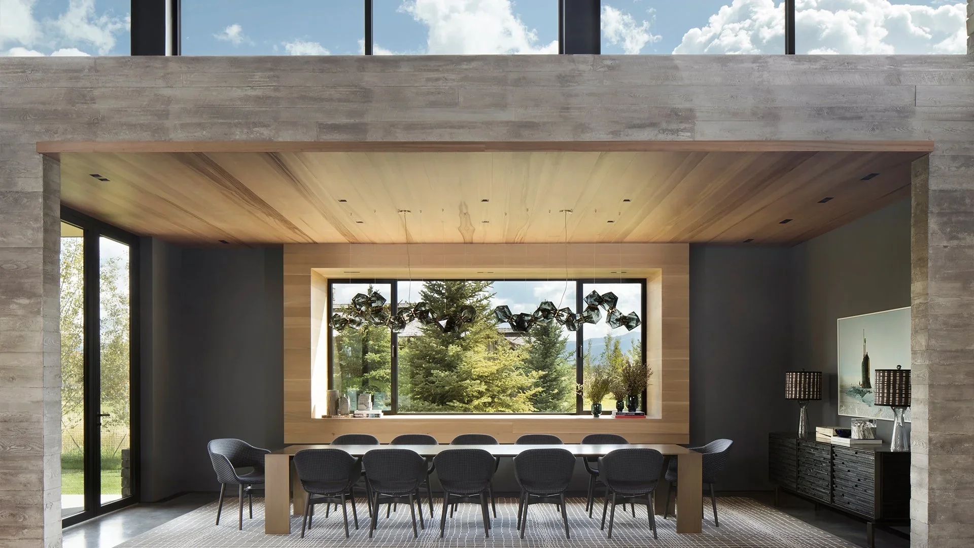 Modern dining room with a long wooden table surrounded by ten black chairs, large window with mountain and trees view, contemporary chandelier, and sideboard with decorative items and lamps.