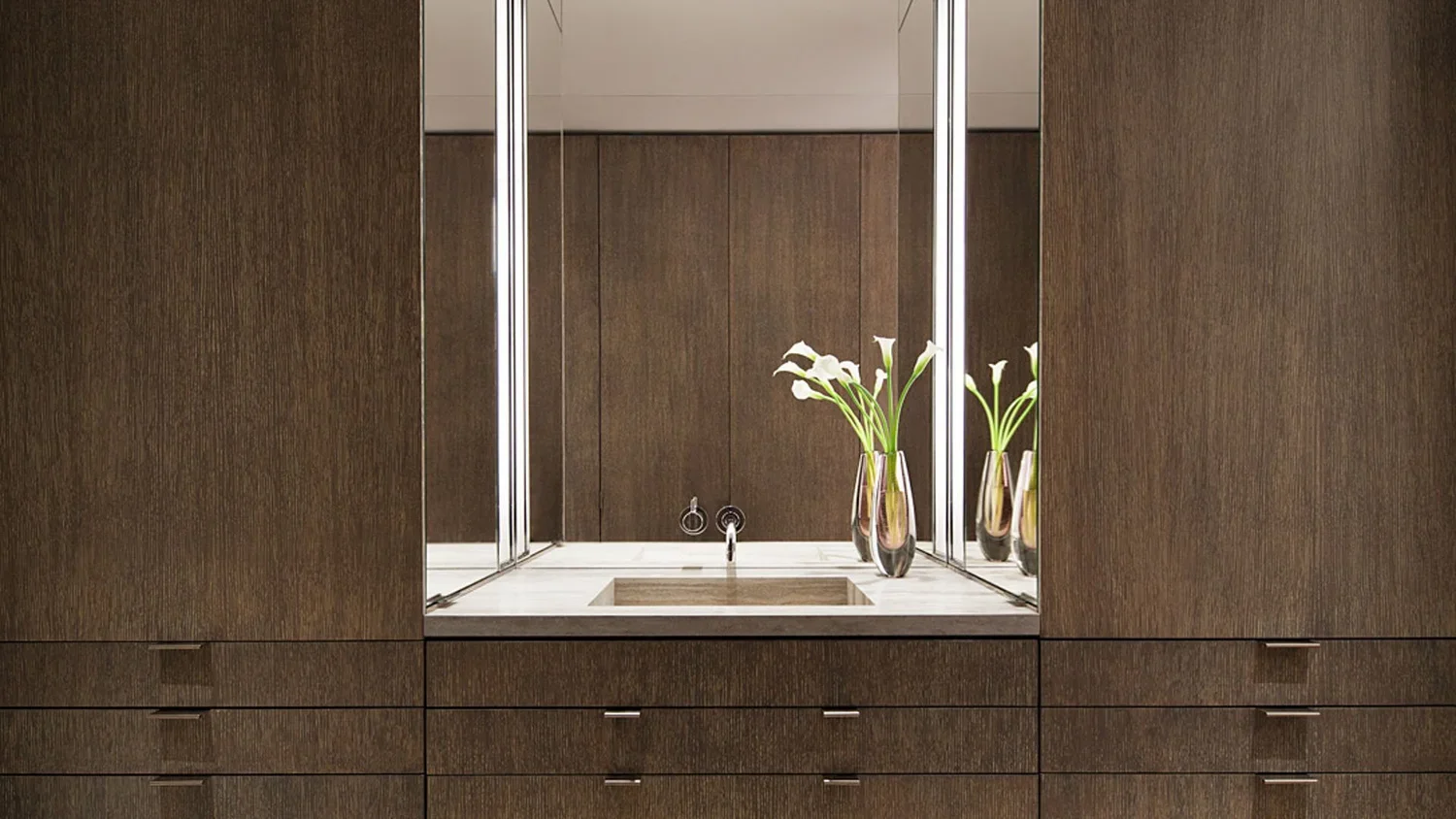 A modern bathroom vanity with a dark wood finish, a white countertop with a built-in sink, a mirror behind, and three vases with white calla lilies on the counter.