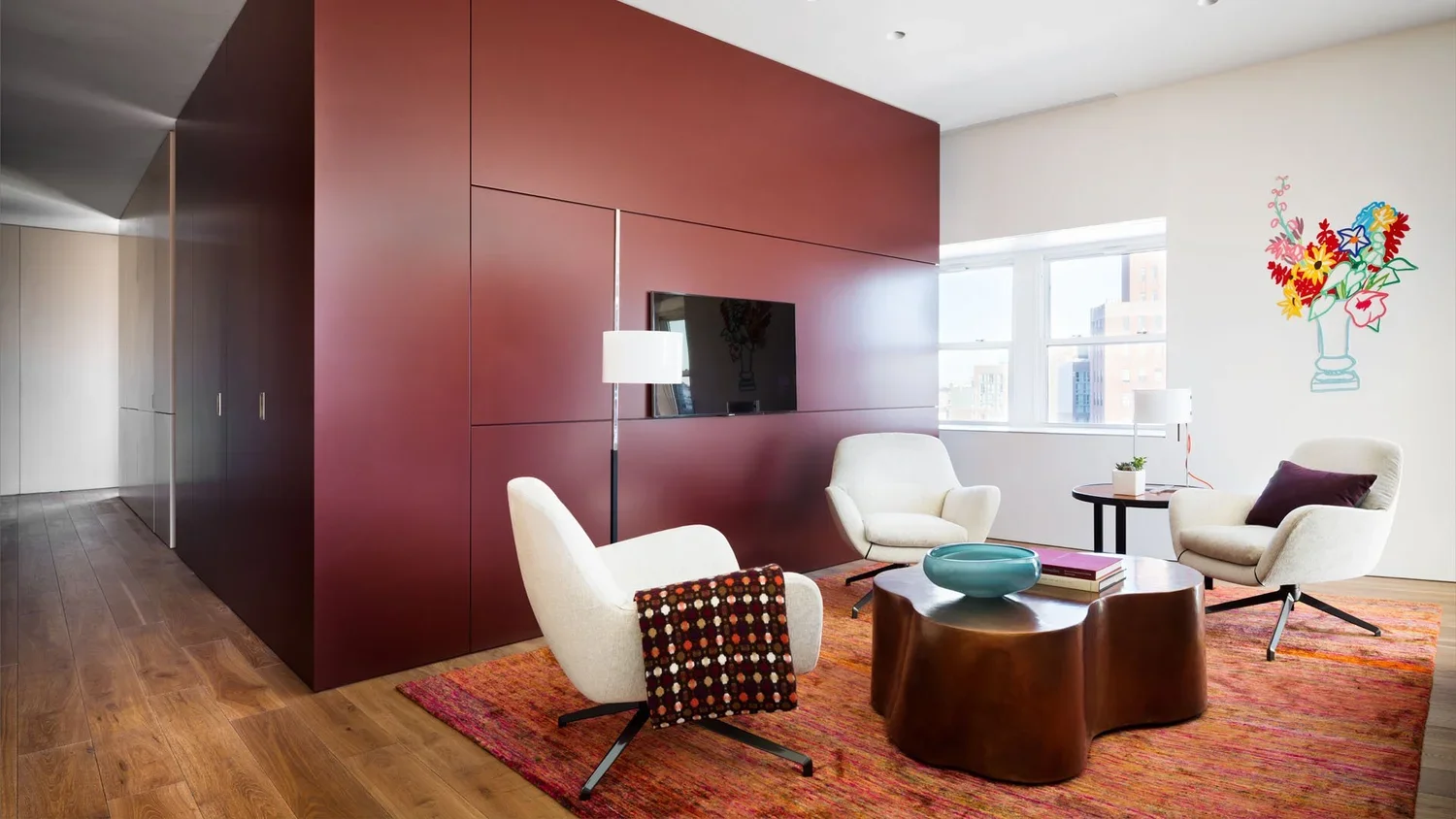 Modern living room with three white armchairs, a round brown coffee table, and a rug on wood flooring. There is a deep red accent wall with a mounted TV, a floor lamp, and a side table with a potted plant. Bright windows and a colorful flower mural d