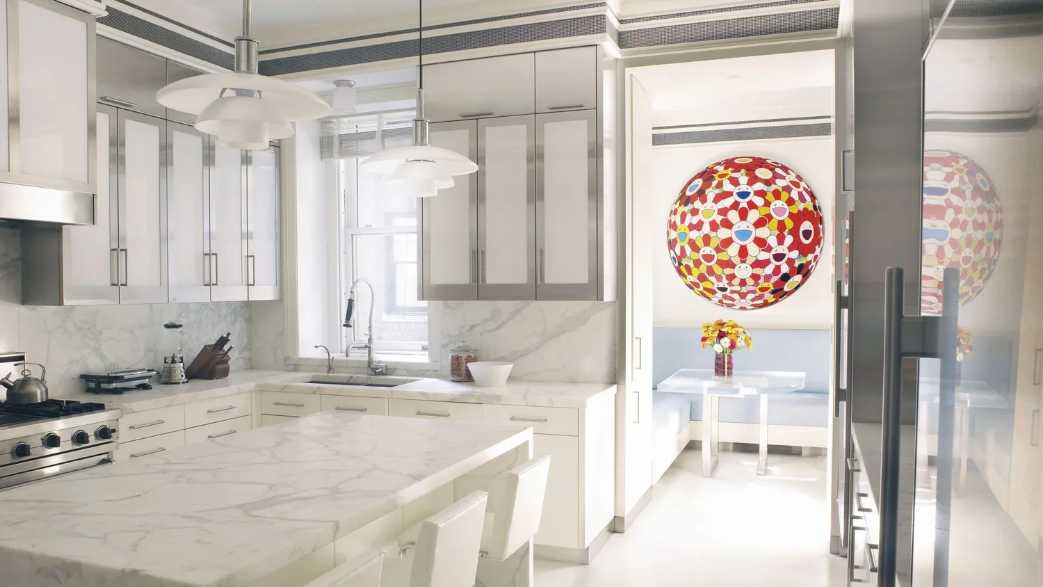 Modern white kitchen with marble countertops, stainless steel appliances, and a brightly colored, patterned spherical sculpture hanging above a dining nook with a window and a table with flowers.
