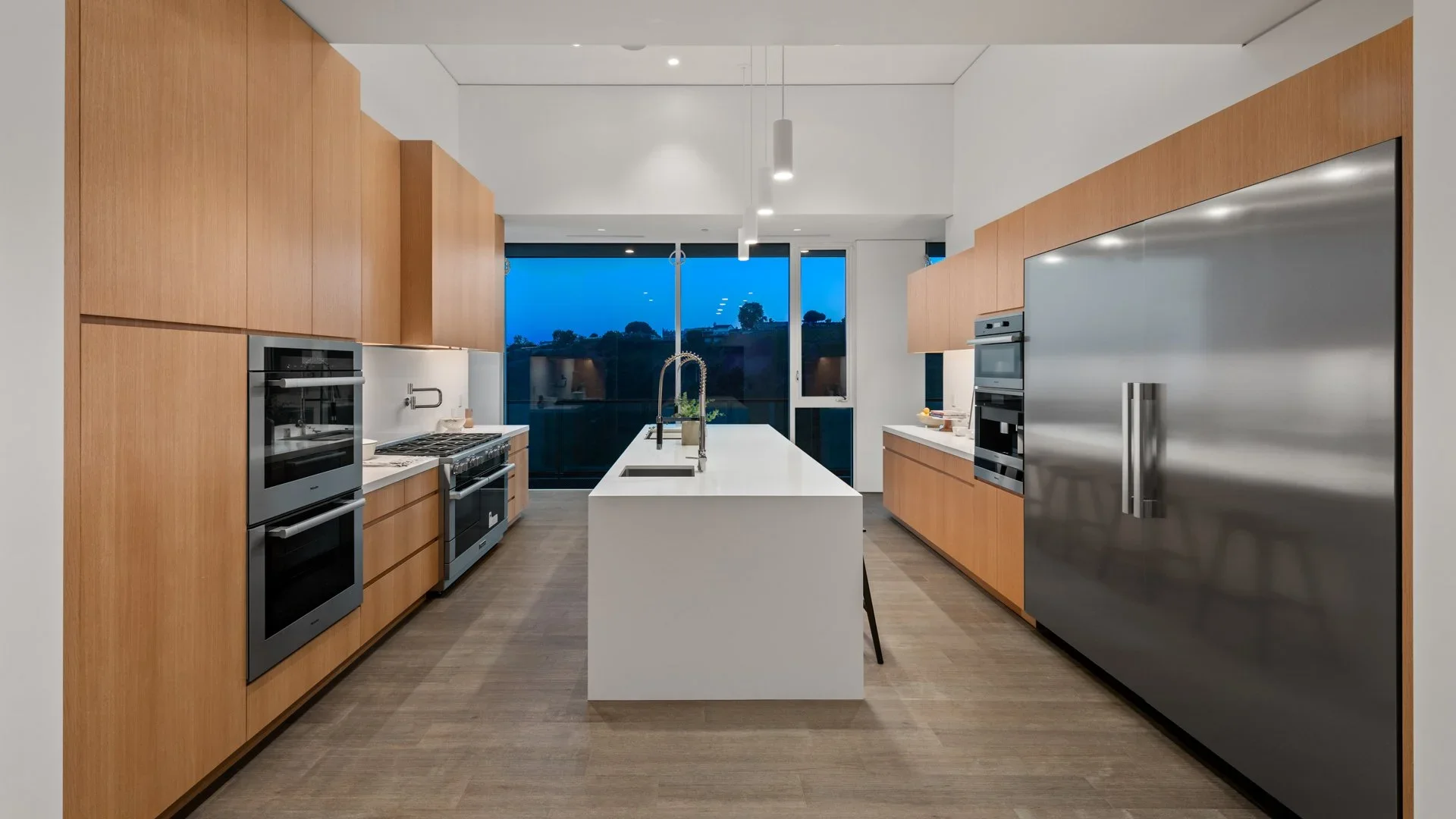 Modern kitchen with wooden cabinets, a white island with a sink, stainless steel appliances, and large windows showing a blue evening sky.