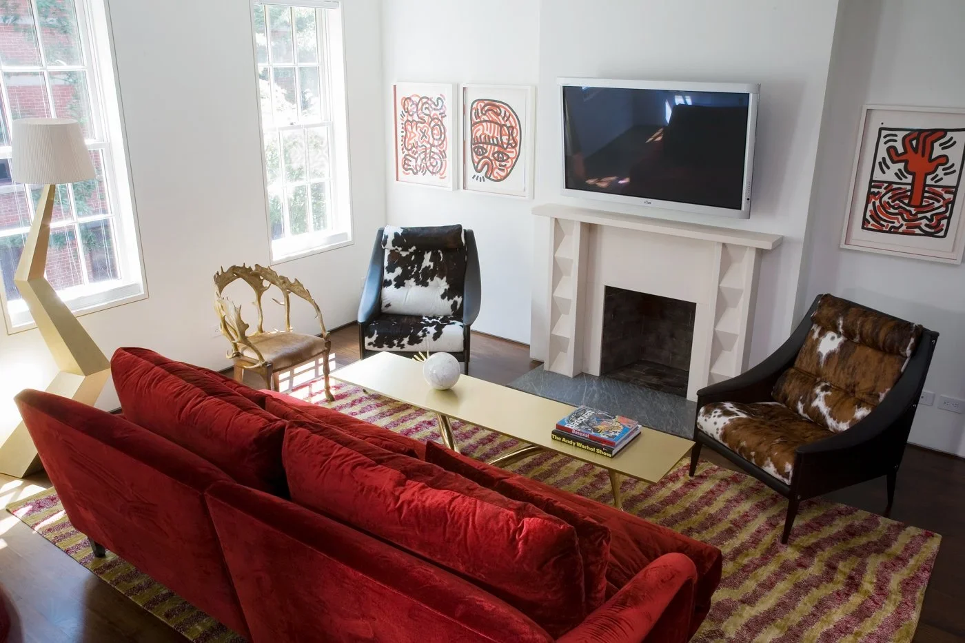 Living room with red velvet sofa, two cowhide armchairs, side chair, yellow coffee table with books, abstract artwork on white walls, large windows, fireplace, television above fireplace, patterned rug, modern floor lamp.