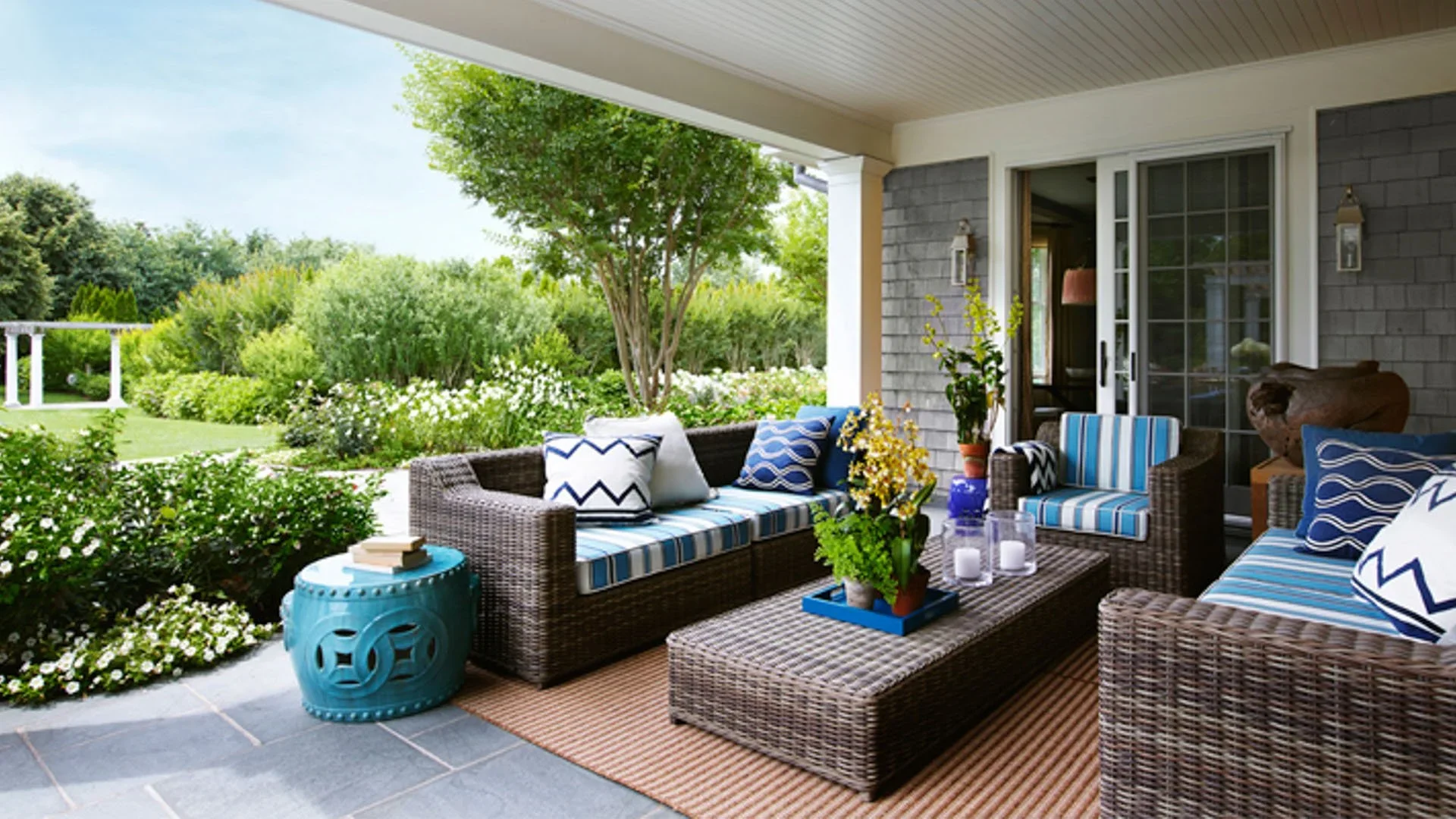 Back porch with wicker furniture, blue striped cushions, and plants, overlooking a green garden with trees and white flowers.