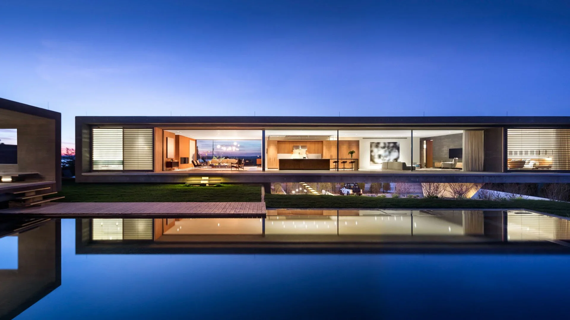 Modern house with large glass windows and interior lighting, reflected in a pool outside during twilight.