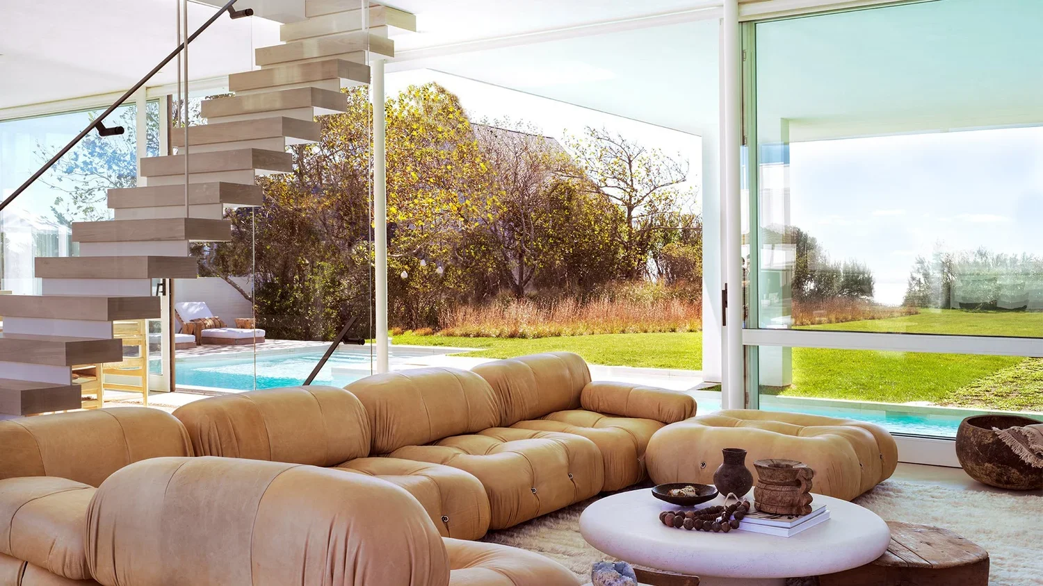 Modern living room with large beige sectional sofa, round coffee table with decorative objects, floor-to-ceiling glass doors overlooking a backyard with trees, grass, and an outdoor pool, modern staircase with open risers.
