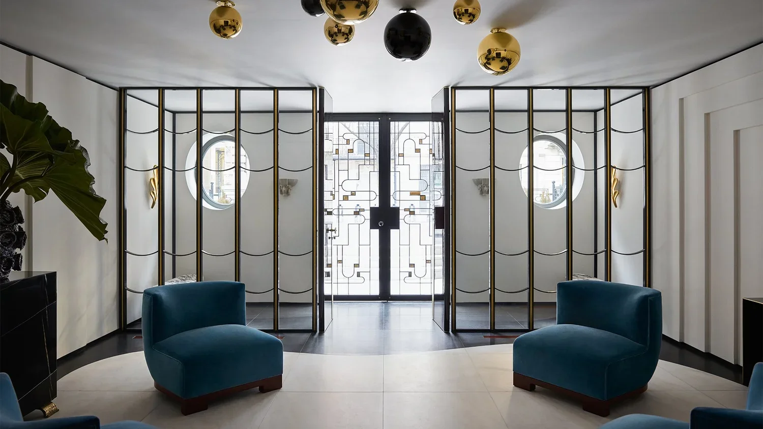 Modern interior hotel lobby with a symmetrical design, featuring two blue armchairs, a glass front door, decorative partitions with circular mirror accents, and gold and black spherical ceiling lights.