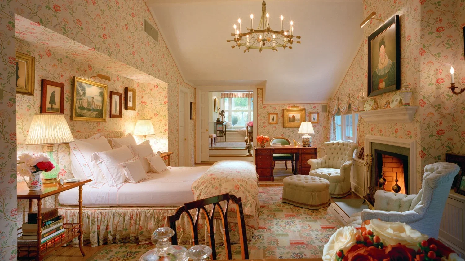 A cozy vintage bedroom with floral wallpaper, a large bed with white bedding, bedside lamps, framed artwork, and a sitting area with armchairs and fireplace.