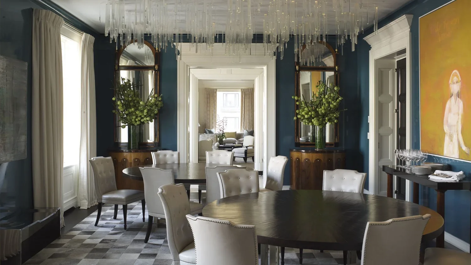 Elegant dining room with round wooden table, white upholstered chairs, blue walls, large vases with green flowers, mirrors, and a crystal chandelier. There is a view into a bright sitting room with large windows.