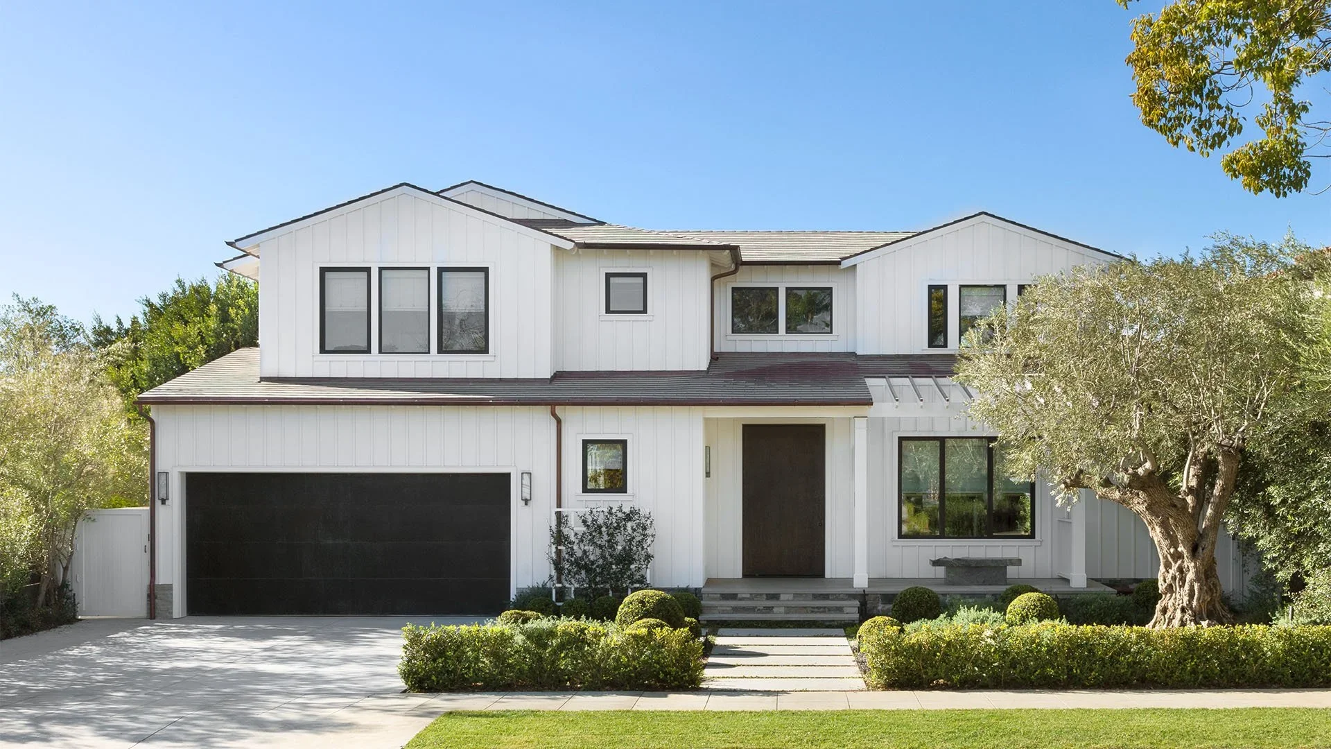 A modern white two-story house with black-framed windows, a black garage door, and a front yard with shrubs, trees, and a concrete pathway leading to the front door.