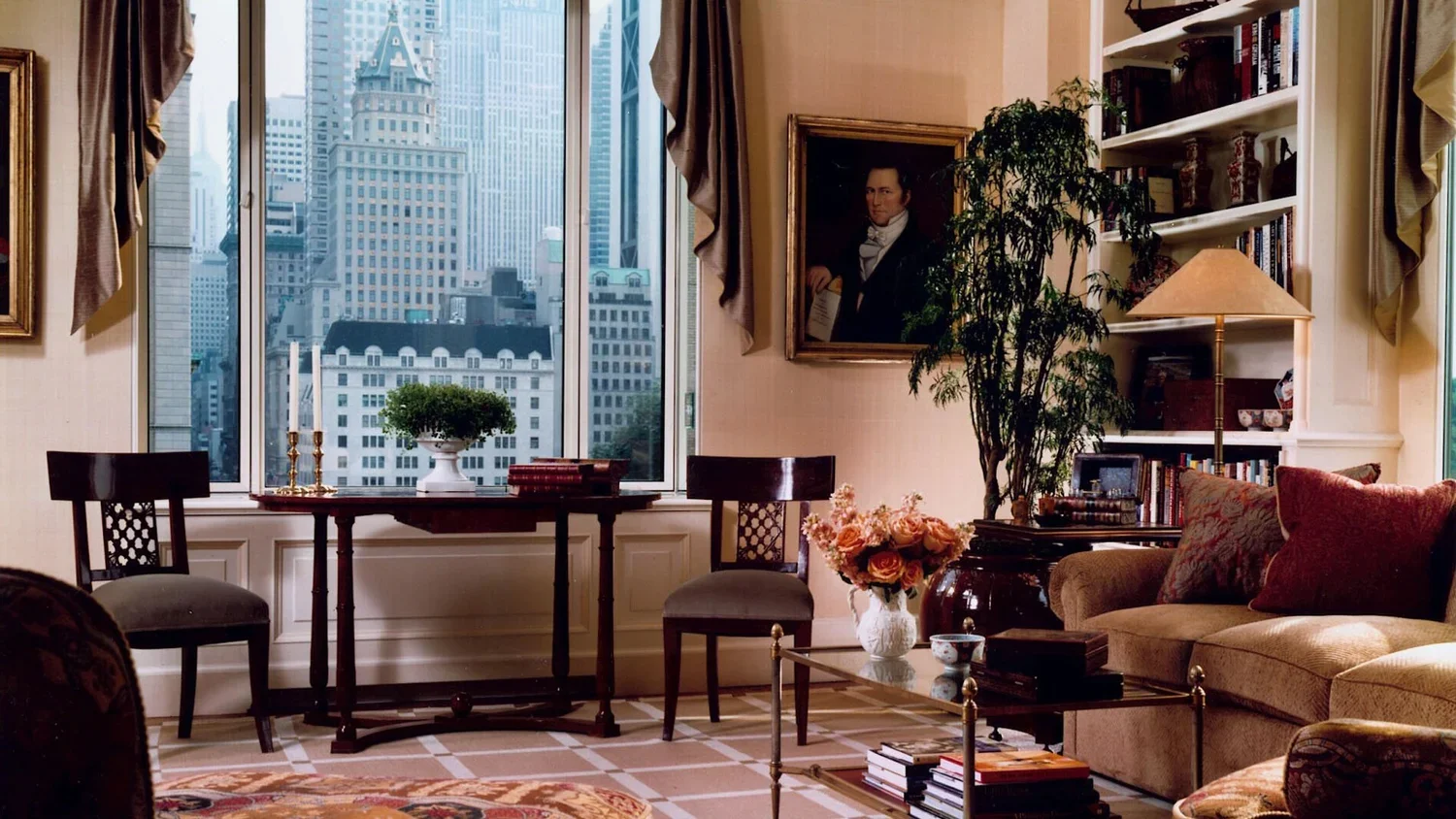 Interior of a living room with large windows showing a cityscape, a sofa with red and brown cushions, a glass coffee table with books and a vase of flowers, a painting on the wall, a tall bookshelf, and a lamp.