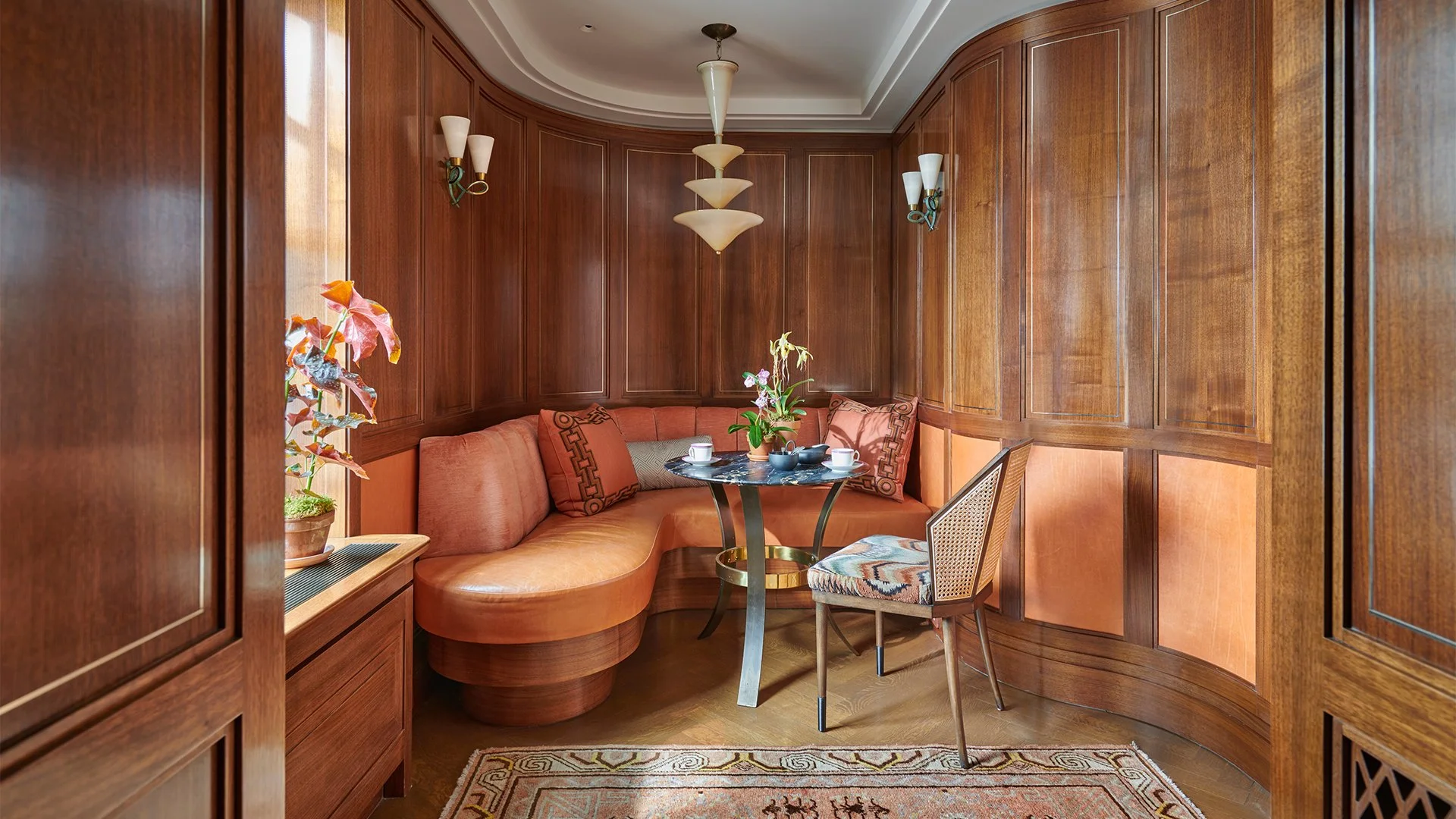 A cozy dining nook with curved wooden panel walls, an orange curved banquette with matching pillows, a round black marble table with a potted orchid and cups, a wooden chair with a patterned cushion, a wall-mounted sconce, a modern multi-tiered penda