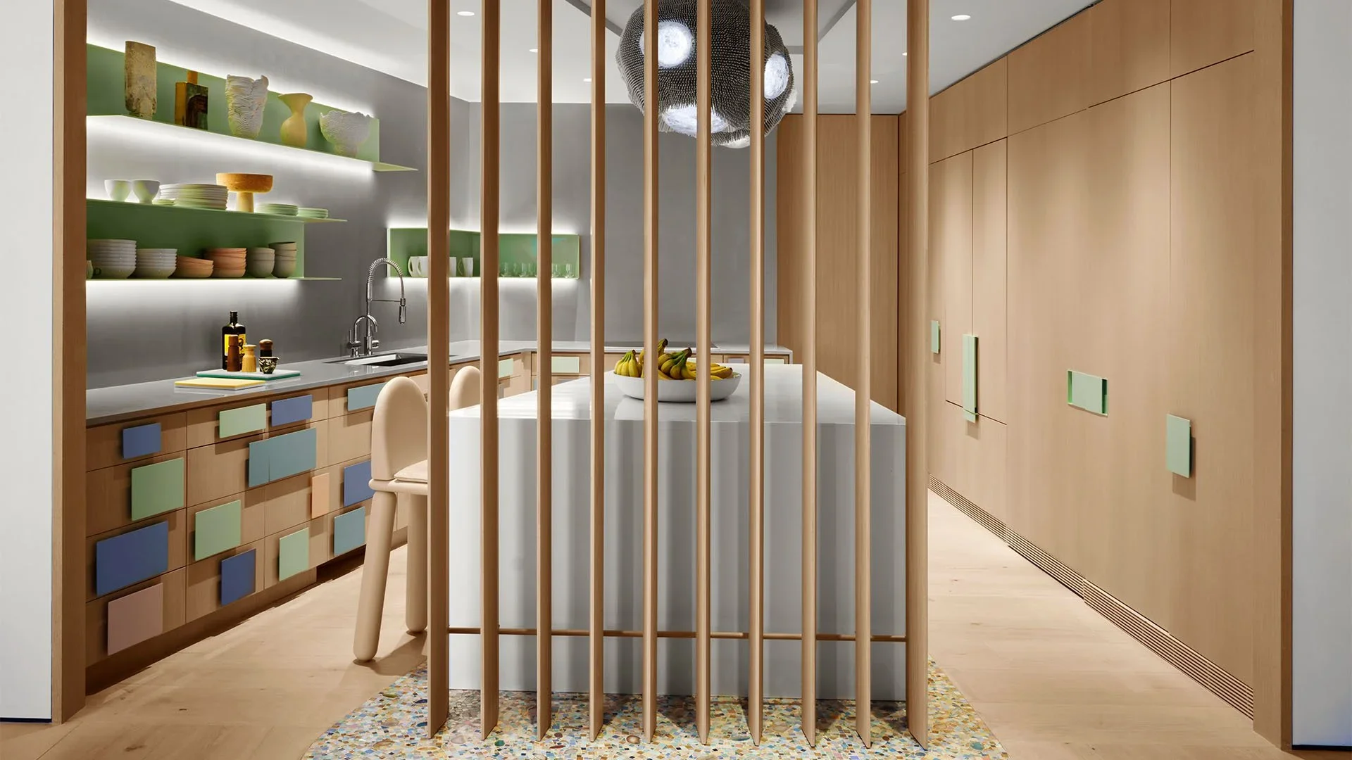 Modern kitchen with light wood cabinetry, open shelves with dishes and vases, a colorful drawer design, and a central island with bananas, separated by wooden slats, and a patterned rug.