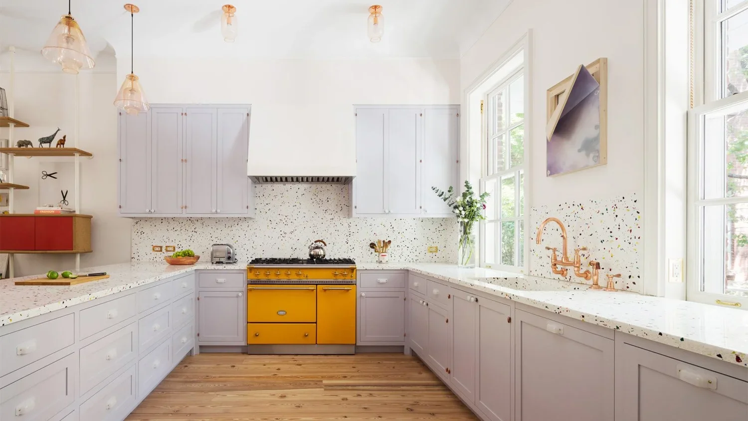 Bright kitchen with white cabinets, terrazzo countertops, and a yellow vintage stove. Open shelves with small decorative items, large windows with view outside, and wood flooring.