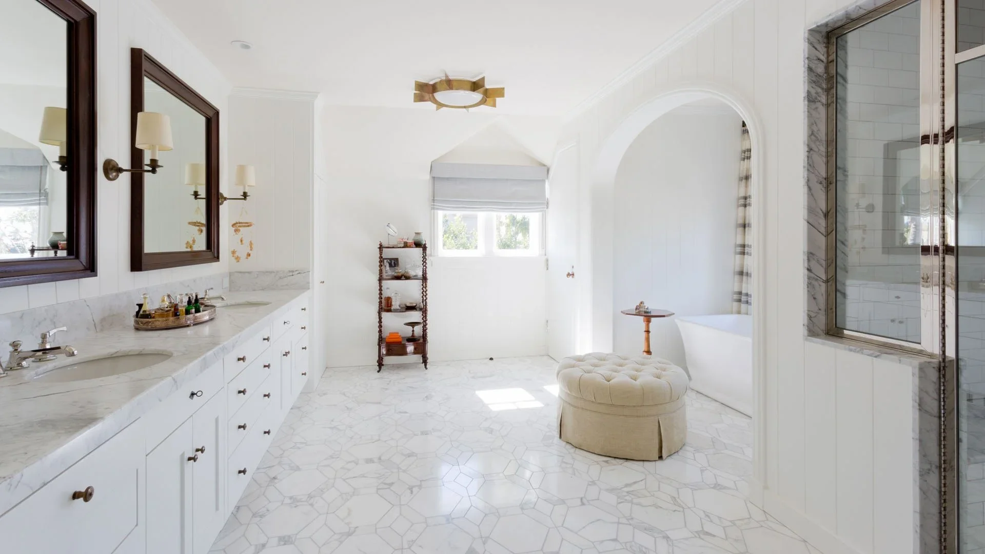 Bright bathroom with white cabinetry, marble counters, double sinks, a tufted round ottoman, bathtub, and large window with shades, in a predominantly white color scheme.