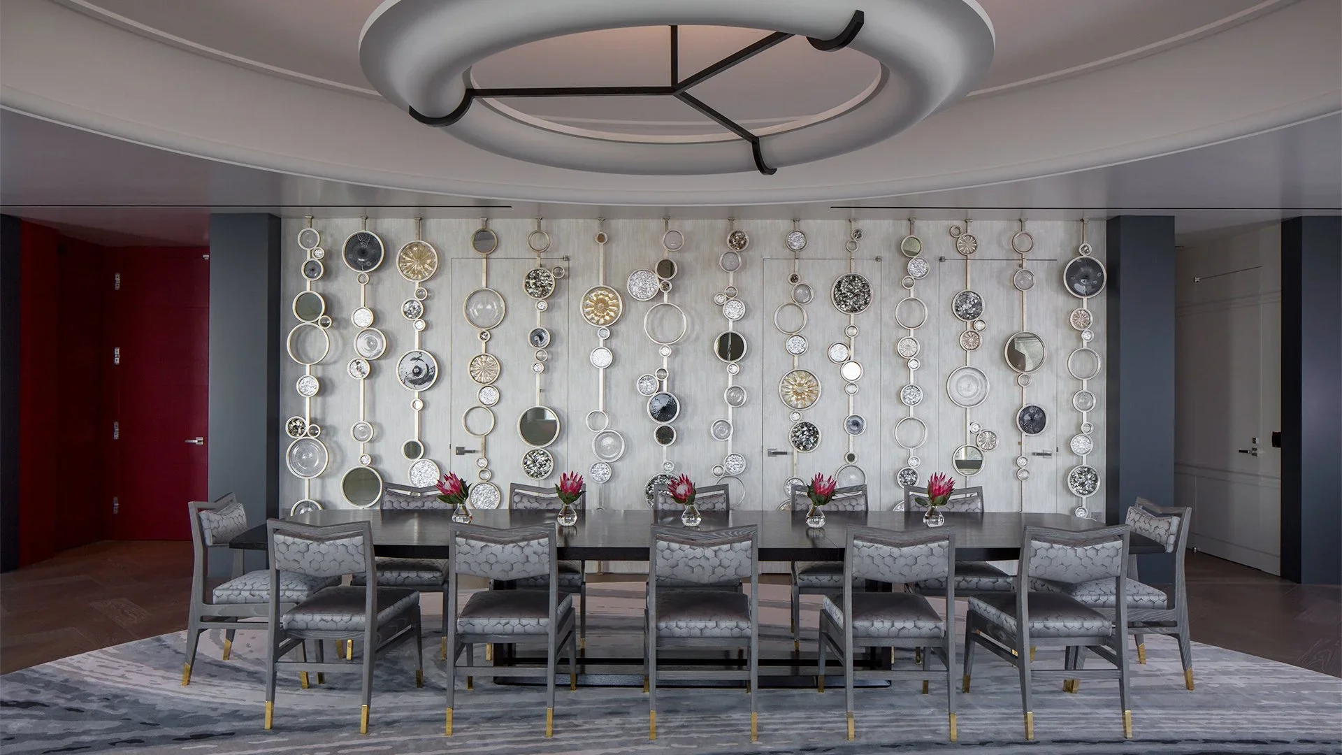 Elegant dining room with a long dark table, 10 upholstered chairs, and pink flowers in glass vases. The wall behind features a decorative pattern of circular mirrors and art pieces.