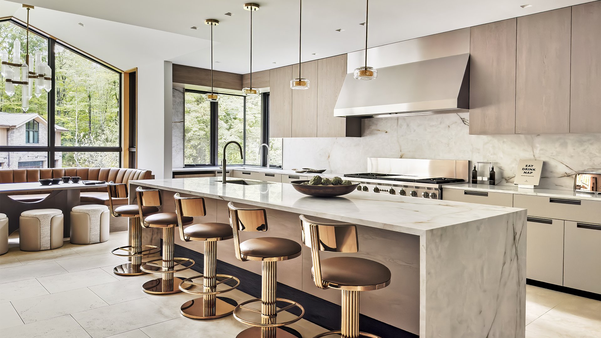 Modern kitchen with large marble island counter, beige and gold bar stools, and a view of green trees through large windows.