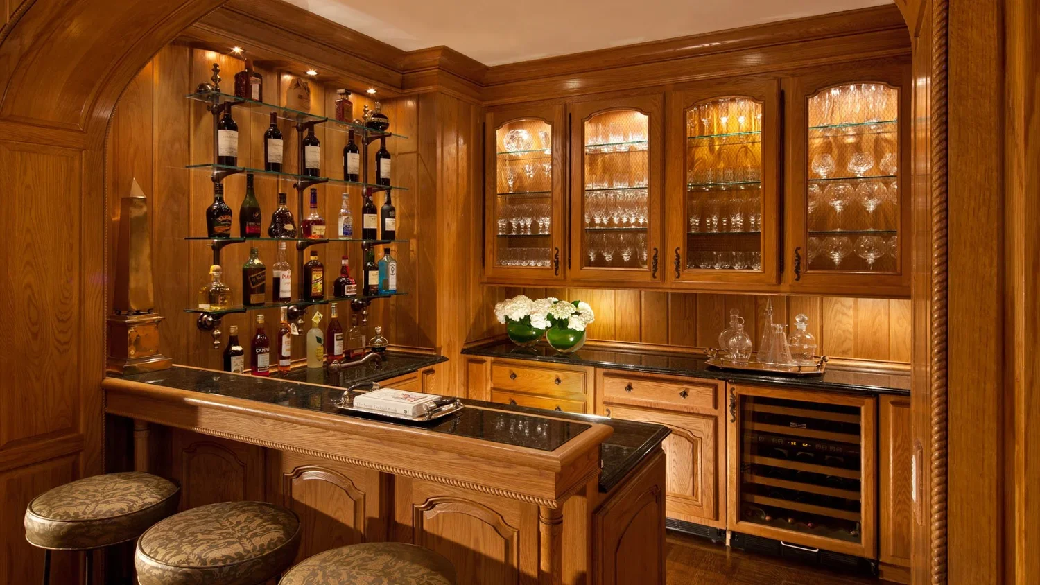 A luxurious home bar with wood paneling, glass-shelved liquor bottles, a black marble countertop, and upholstered barstools.