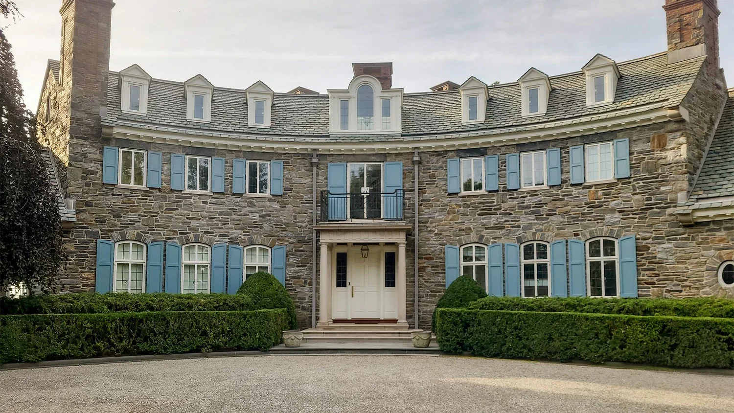 Large stone mansion with blue shutters, multiple windows, and a centered front door with a small balcony above it. Well-manicured hedges and a gravel driveway in front.