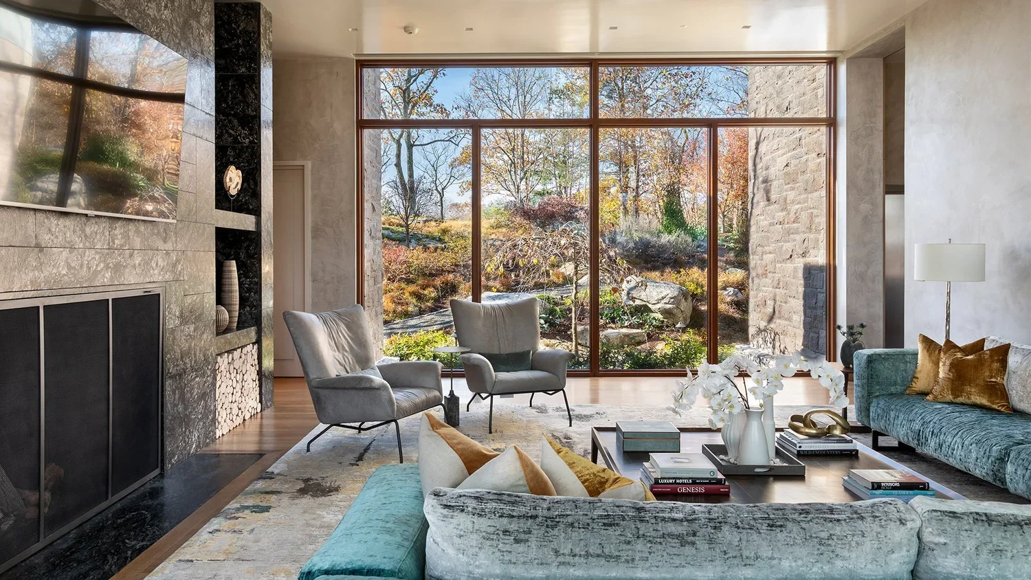 Modern living room with large floor-to-ceiling windows overlooking a landscaped garden with trees and rocks, furnished with a turquoise velvet sofa, gray armchairs, a wooden coffee table, and a fireplace with decorative shelves.